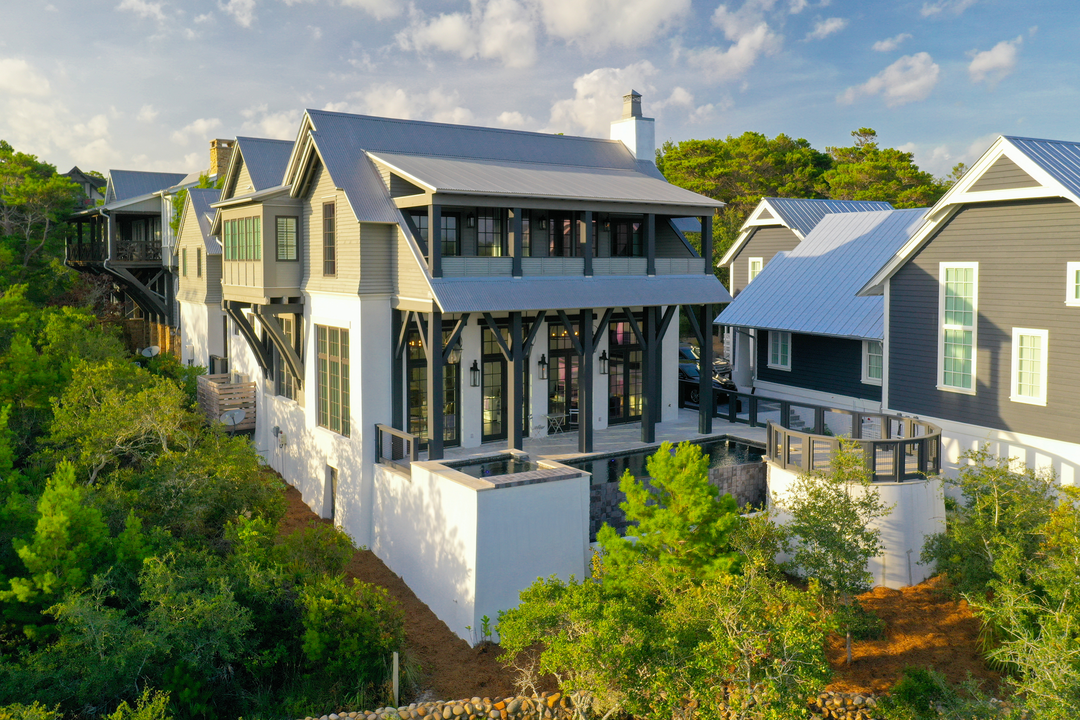 A modern multi-story house with large glass windows, black and white exterior, surrounded by greenery and trees, under a partly cloudy sky.
