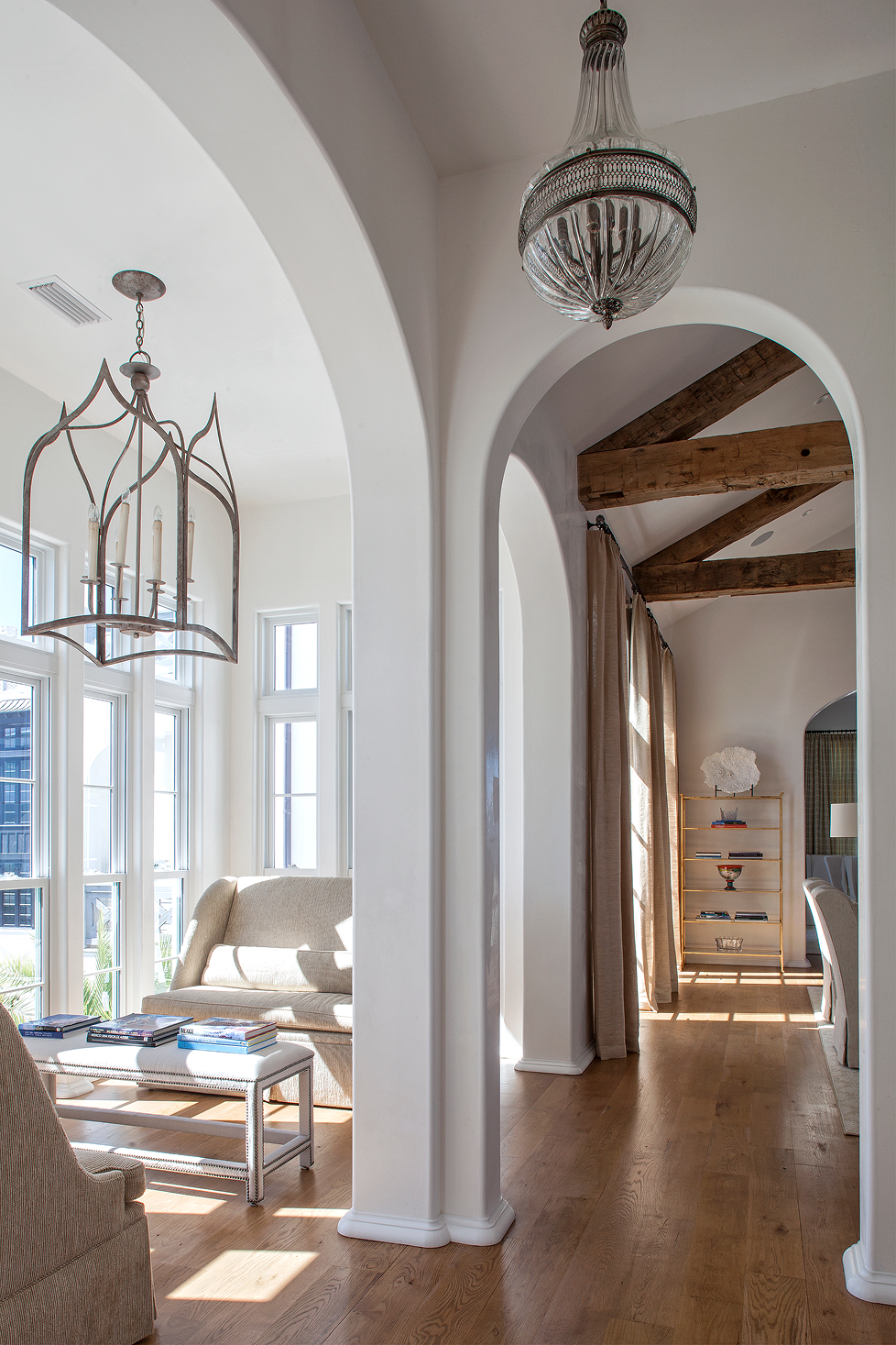 A bright living room with large windows, light-colored furniture, and wooden ceiling beams, visible through two arched doorways.