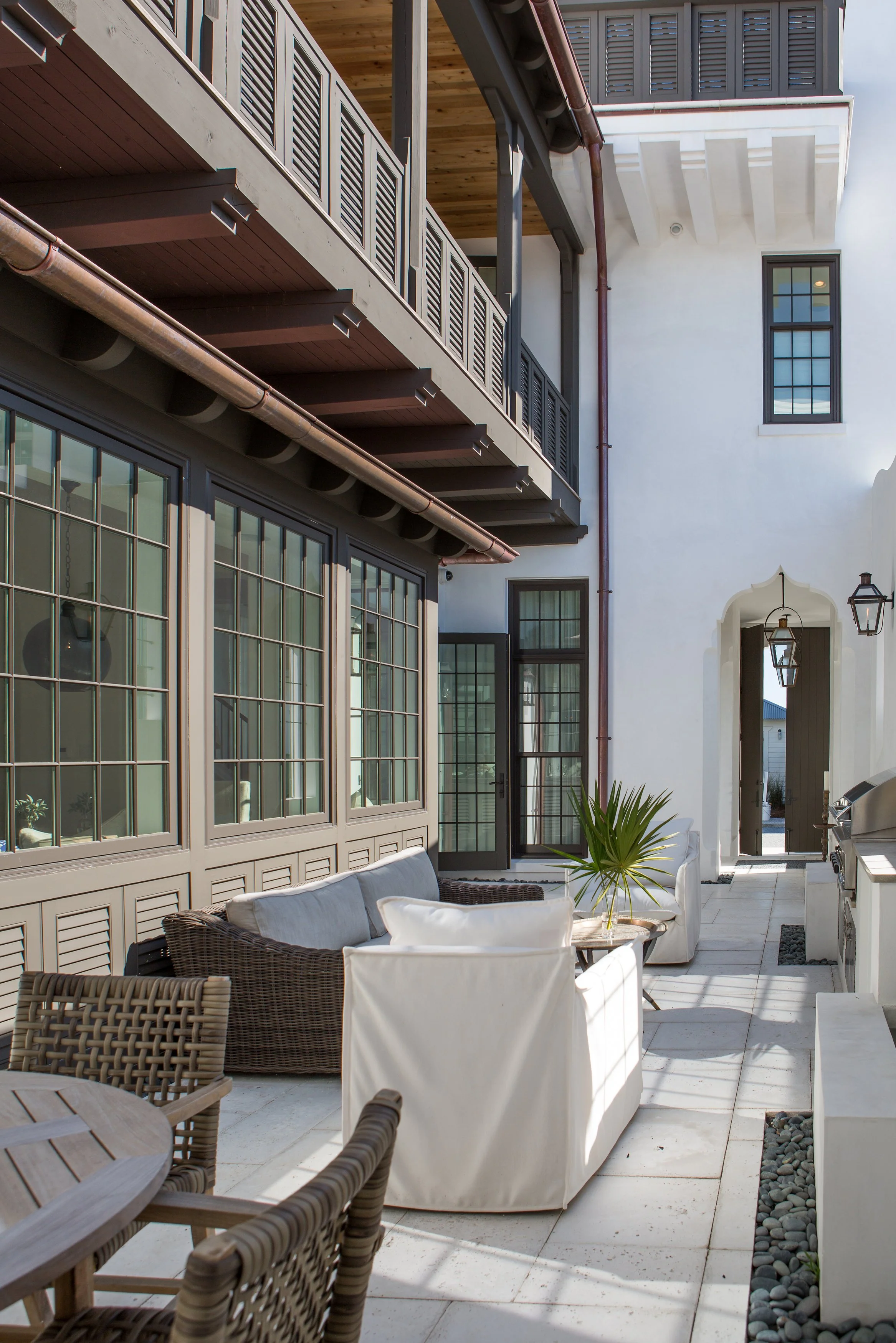 Outdoor patio area with wicker and upholstered seating, a table with green plant, large windows, and an open doorway leading inside a modern building.