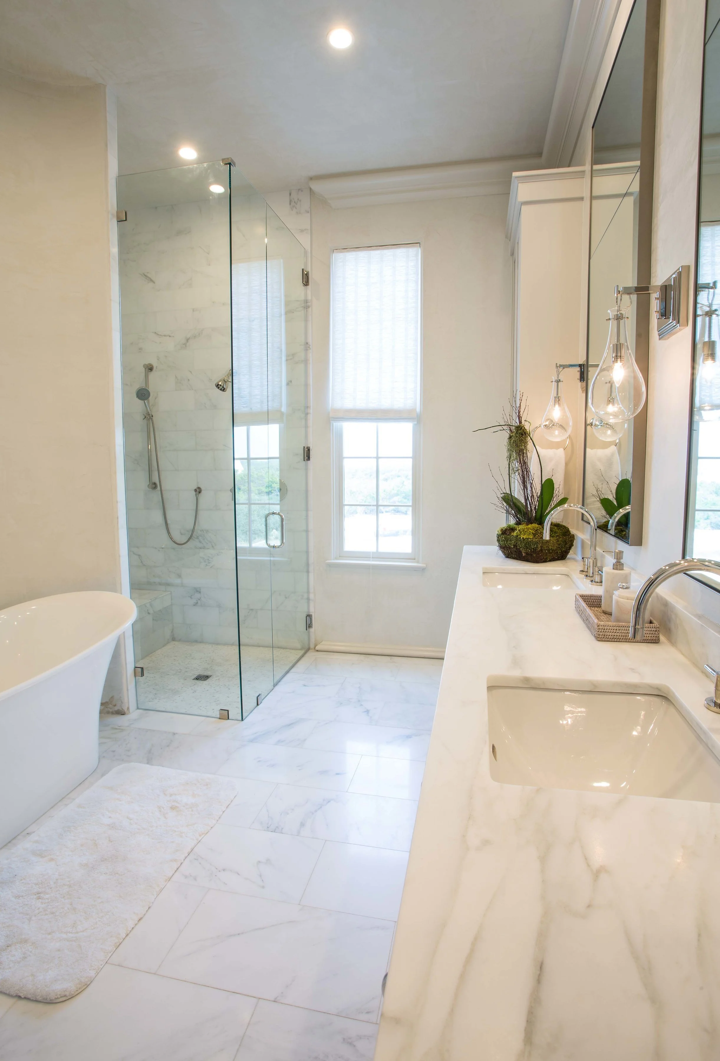 A luxurious bathroom with a white marble vanity, a glass-enclosed shower, a freestanding bathtub, and a window with light-colored blinds.