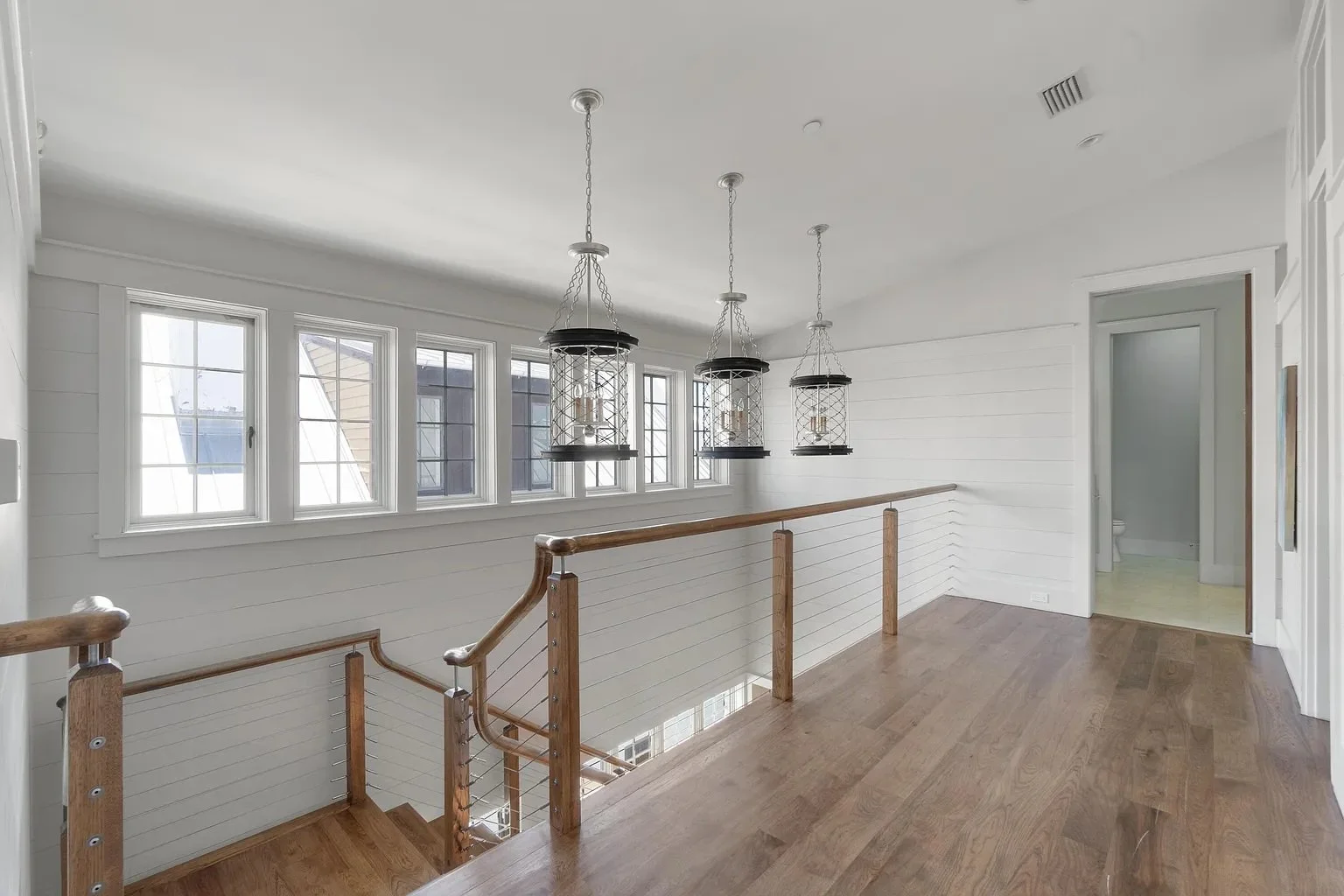 View of a bright second-floor landing with white shiplap walls, four windows, three black lantern pendant lights, wooden handrail and flooring, and a doorway to a bathroom.