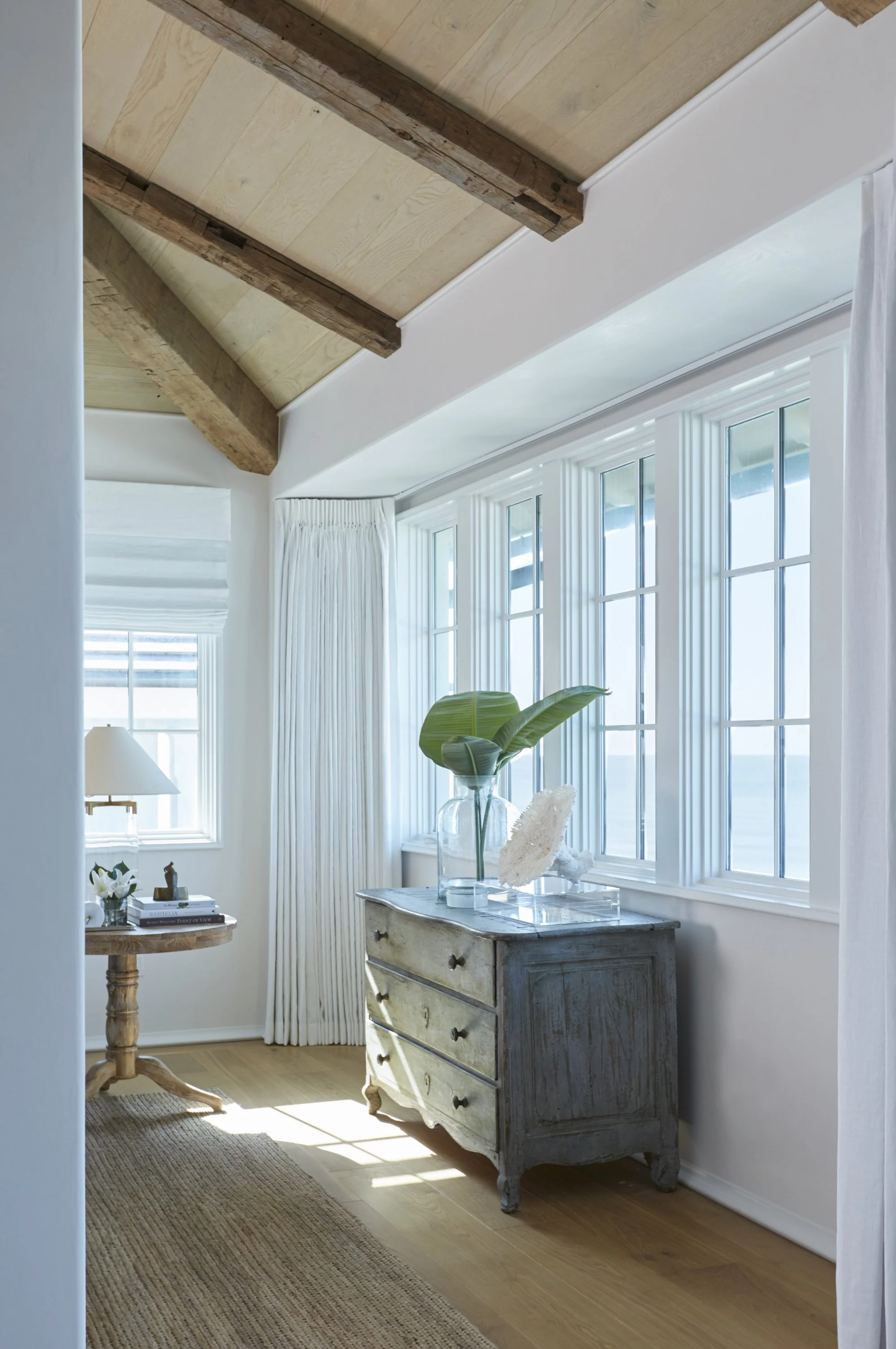 Bright interior room with large windows, white curtains, a vintage gray dresser with decorative items, a houseplant, and a wooden ceiling with beams.