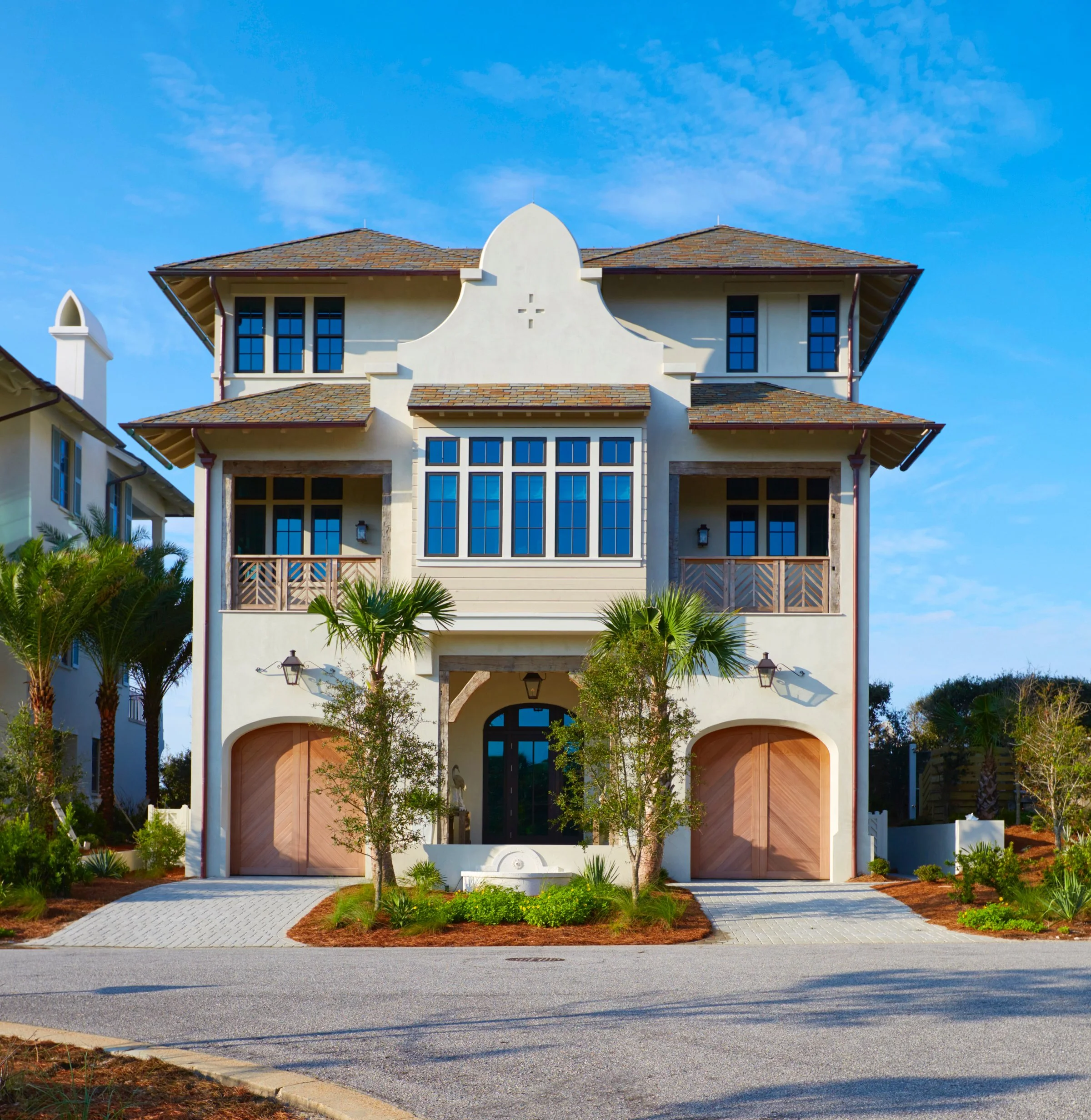A large three-story house with a beige stucco exterior, blue windows, and a mansard roof with brown shingles. The house has two garage doors, a main entrance with a black door, and surrounding greenery including palm trees and landscaped bushes.