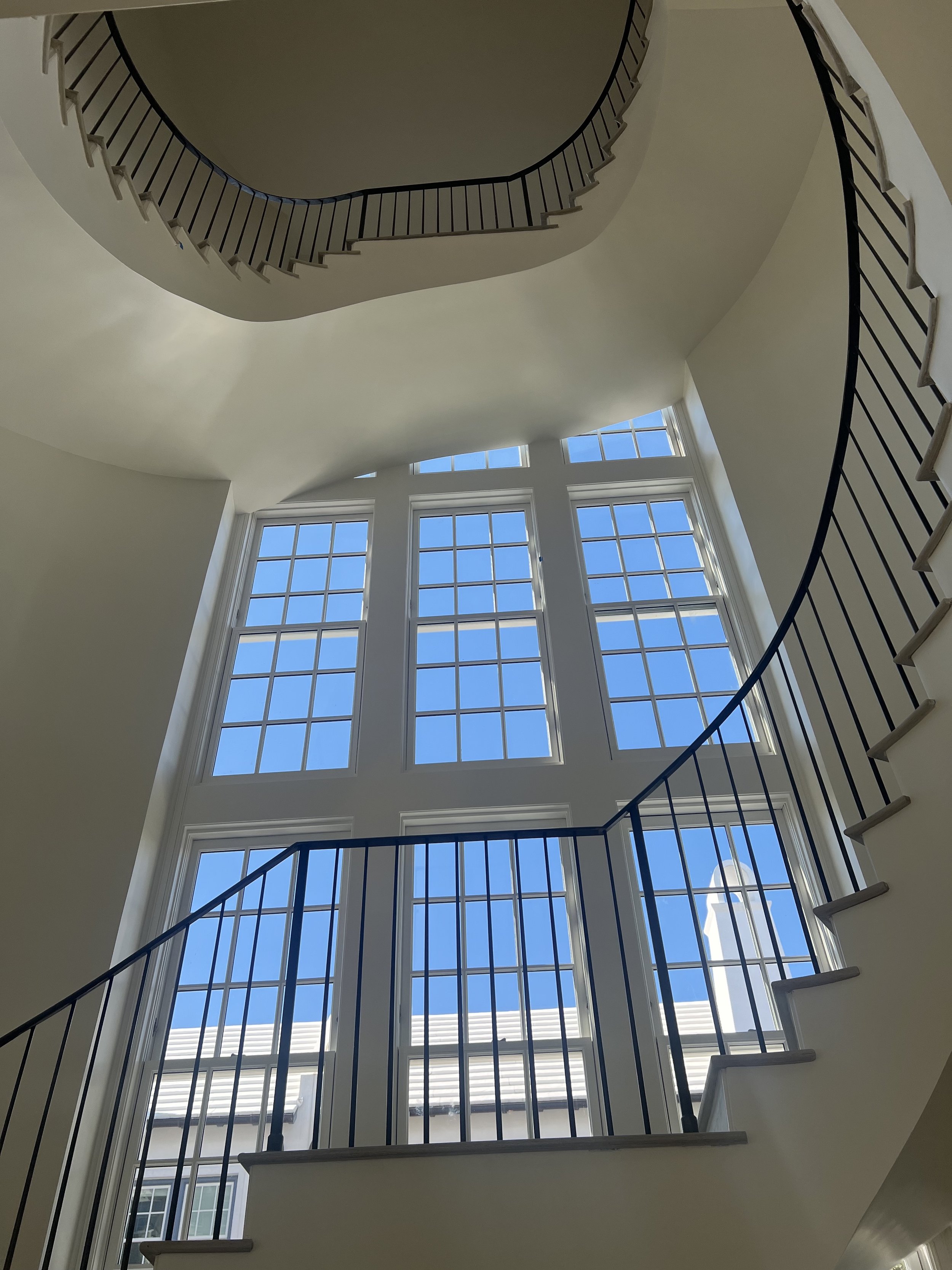 Interior view of a staircase with black metal railings, large multi-pane windows showing a bright blue sky outside, and a curved architectural ceiling.
