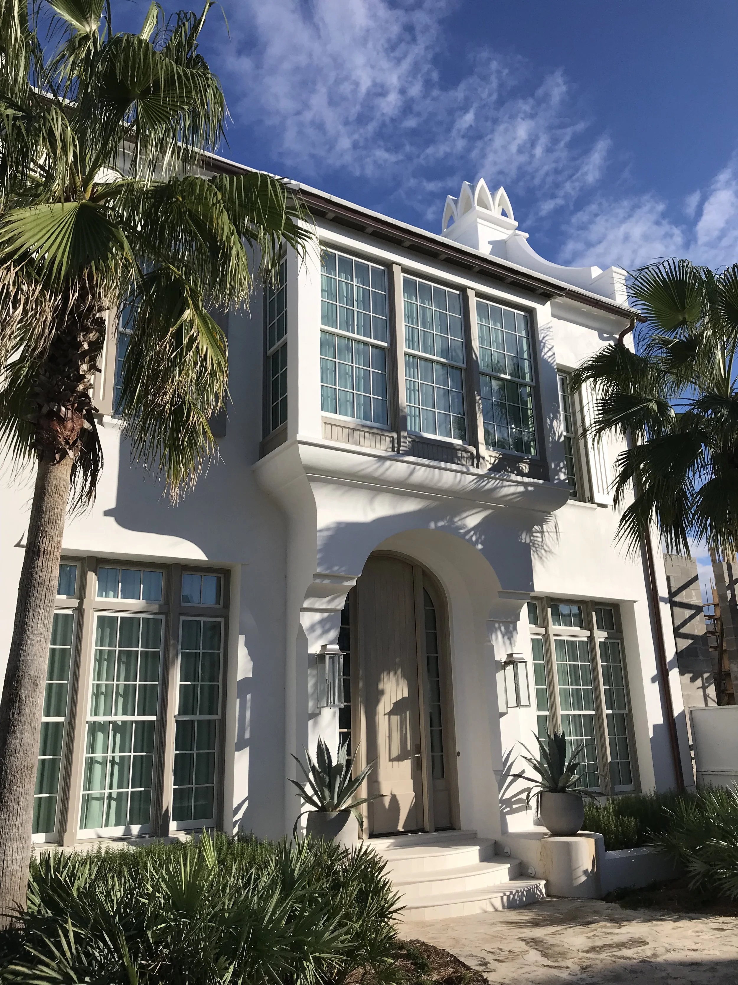 A white modern house with large windows, two potted plants at the entrance, palm trees, and a blue sky with clouds.