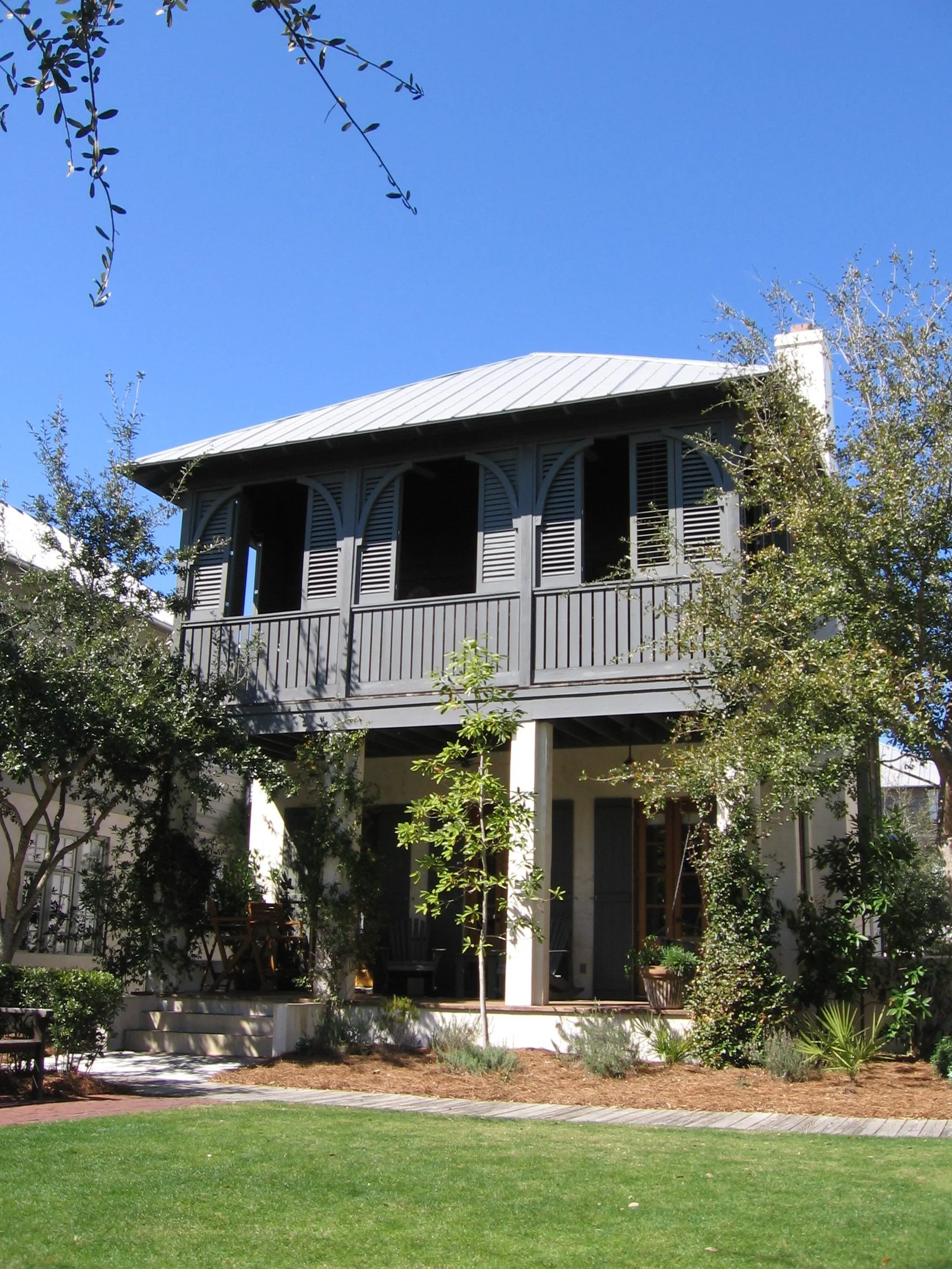 A two-story house with a dark gray exterior, a metal roof, and a balcony on the second floor. The house is surrounded by trees and a well-maintained lawn.