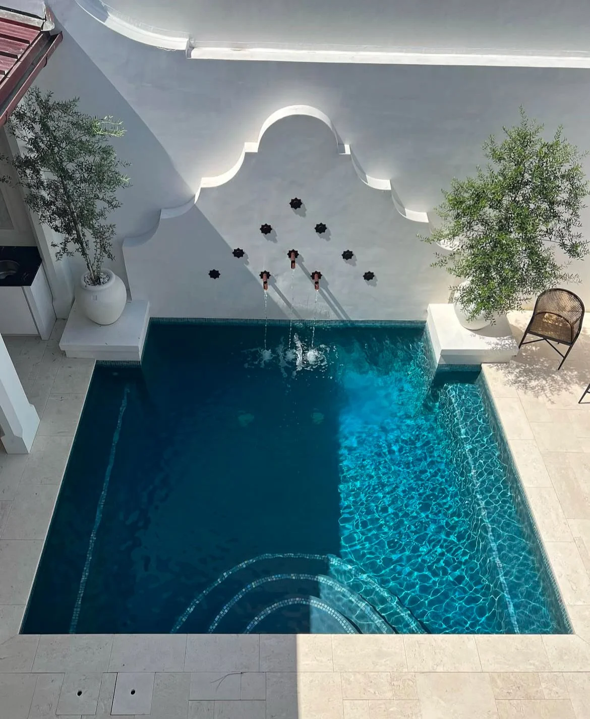 Top-down view of a swimming pool with water features and a decorative wall with arches, flanked by two potted trees and a chair.