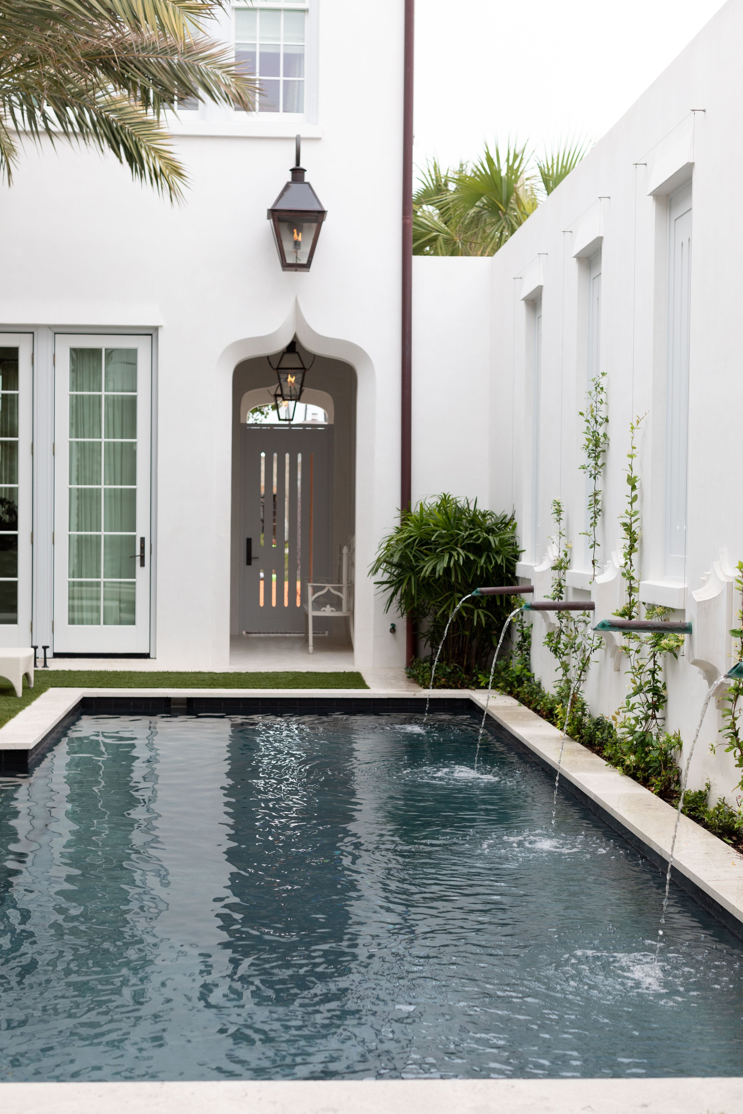 A small rectangular pool with dark water is situated in a courtyard, surrounded by white walls. There are three water spouts along the right wall, guided by pipes, and a fountain in the middle. Green plants grow along the wall, and a sliding glass do
