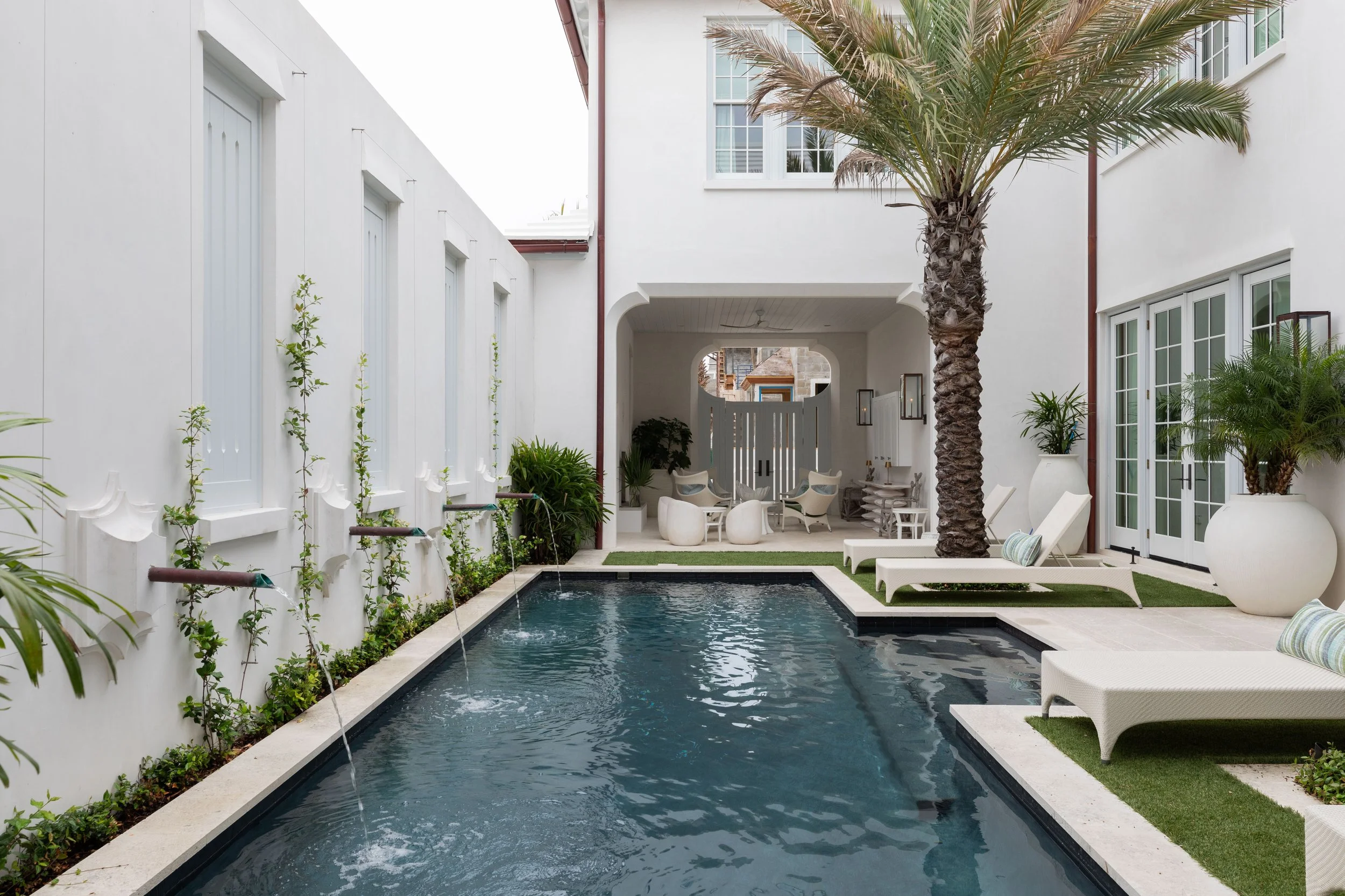 A modern outdoor pool area with a rectangular pool, surrounded by white walls, palm trees, lounge chairs, and potted plants, adjacent to a white building.