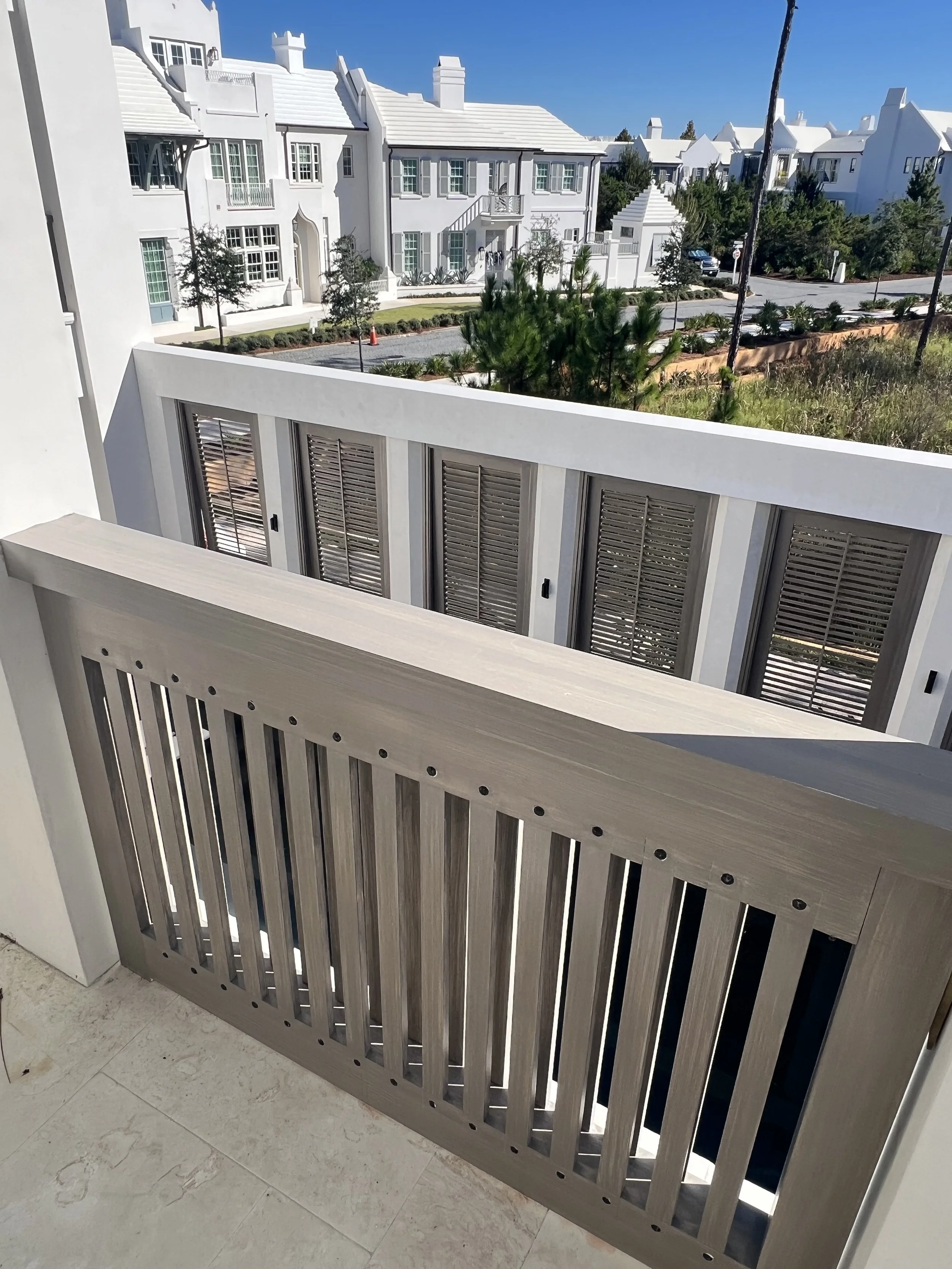 View of a residential neighborhood with white houses, trees, and a clear blue sky. The photo is taken from a balcony with a wooden railing.