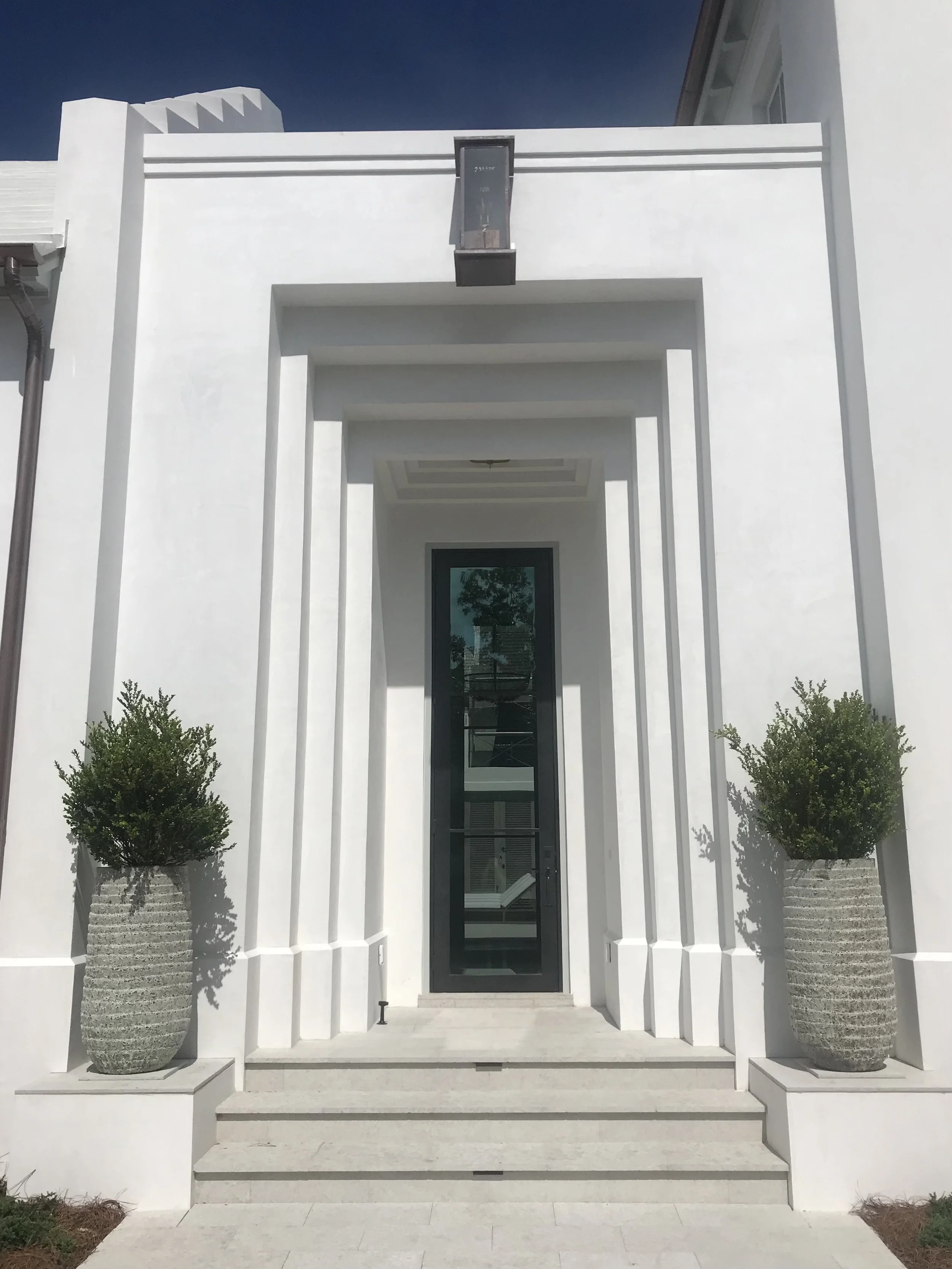 Modern white building entrance with steps, a black door, two potted plants, and decorative architectural elements.