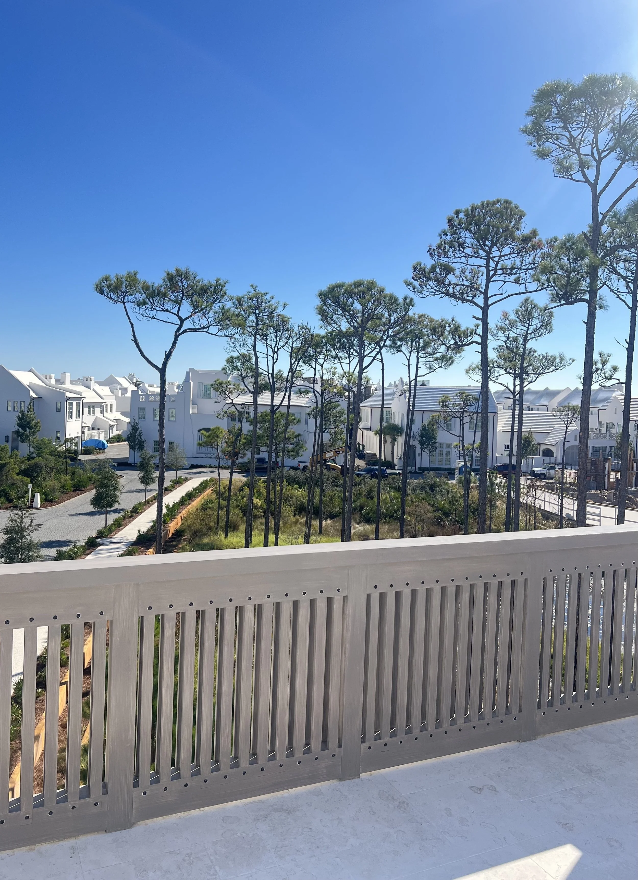 A view from a balcony with a beige wooden railing, overlooking a neighborhood with white houses, trees, and a clear blue sky.