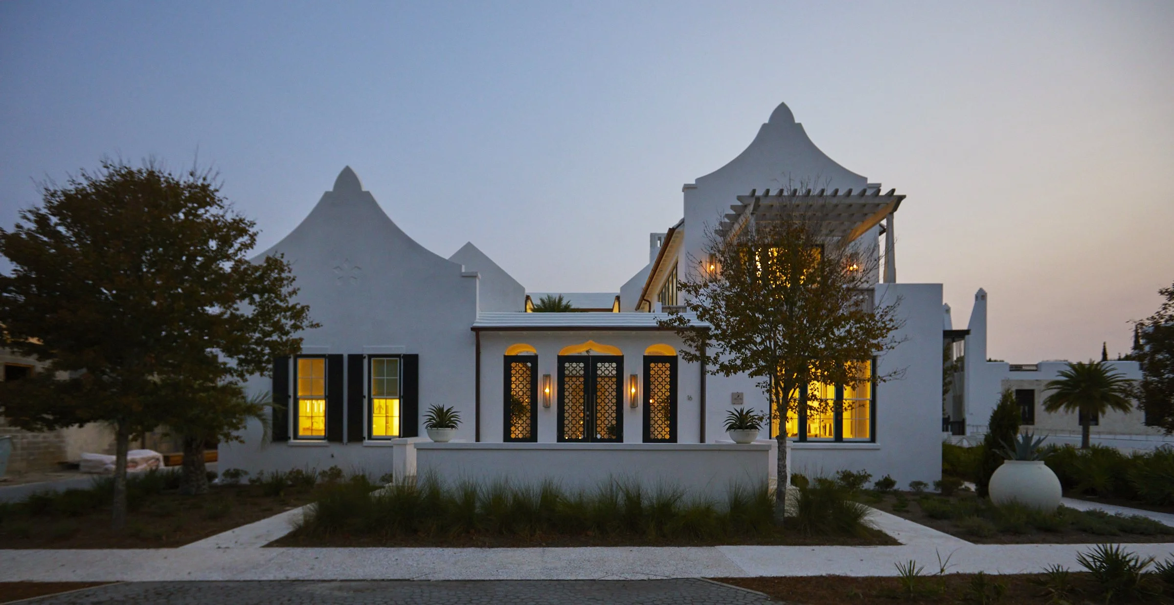 White modern house with black window shutters and a decorative facade, illuminated from inside, surrounded by landscaped garden with trees and potted plants, during dusk or dawn.