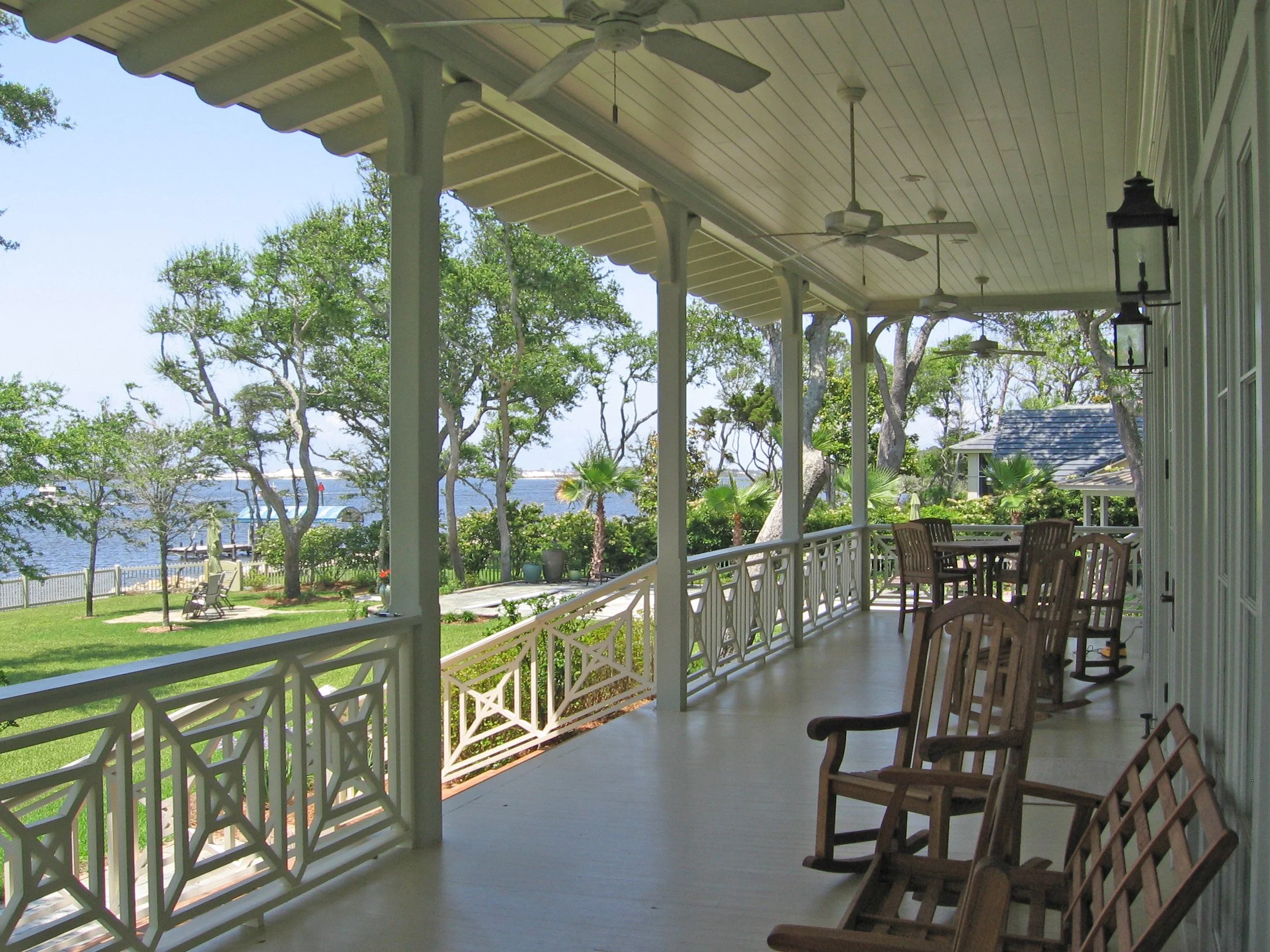 View from a porch overlooking a backyard with grass, trees, and water in the distance. The porch has wooden chairs, ceiling fans, and lantern-style light fixtures.