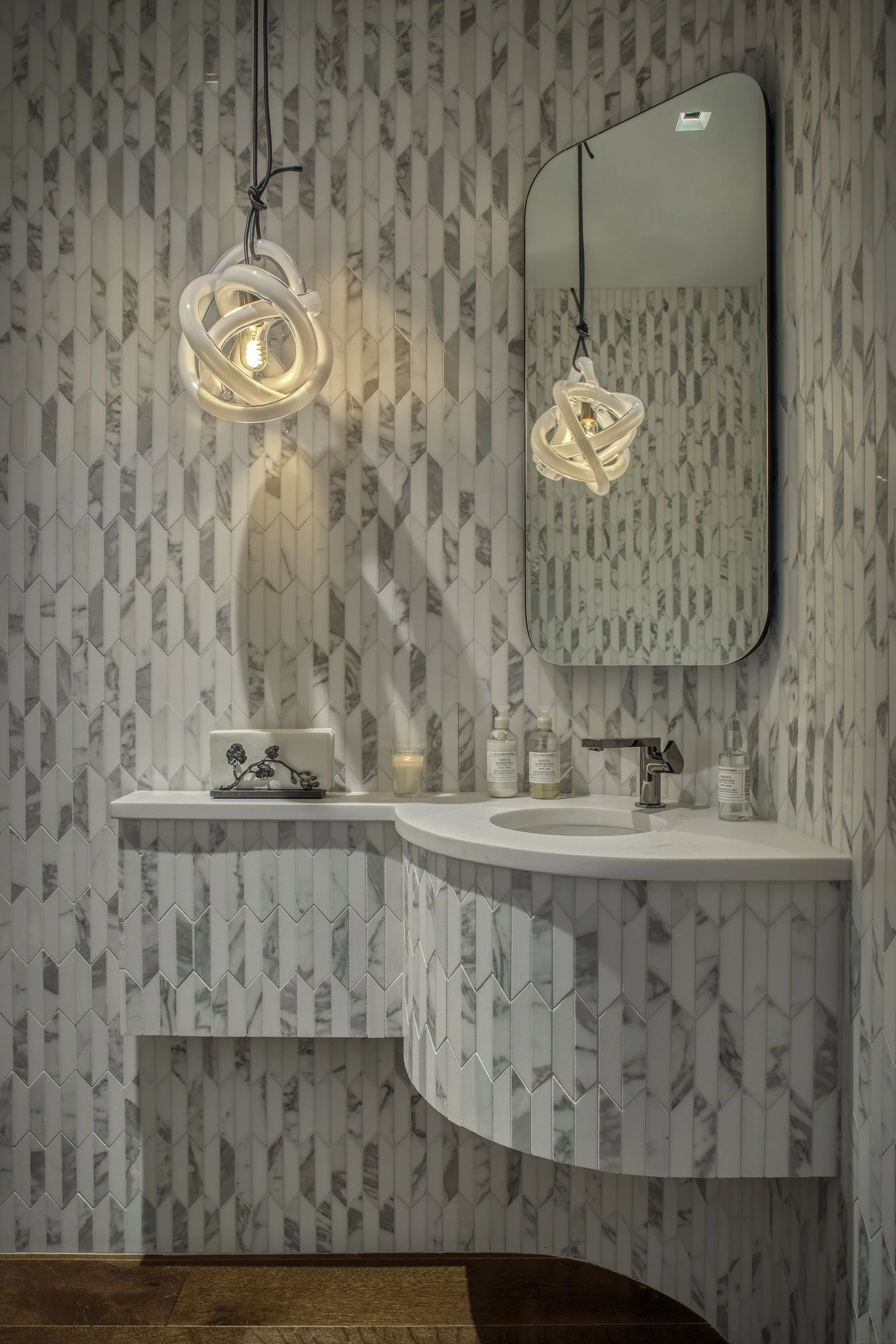 A modern bathroom with a marble-tiled wall, a curved marble sink, an oval mirror, and a distinctive sculptural light fixture.