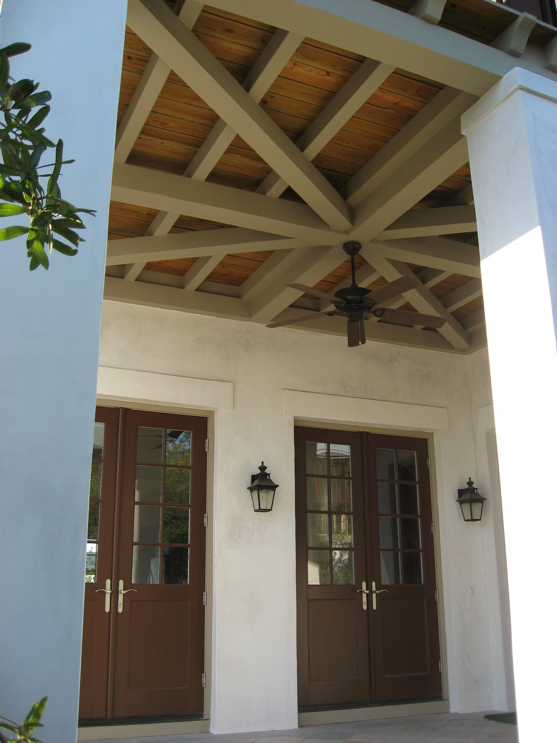 View of a covered porch with two brown French doors, black wall-mounted lanterns on each side, and a ceiling fan with wooden blades under a wooden-beamed ceiling.