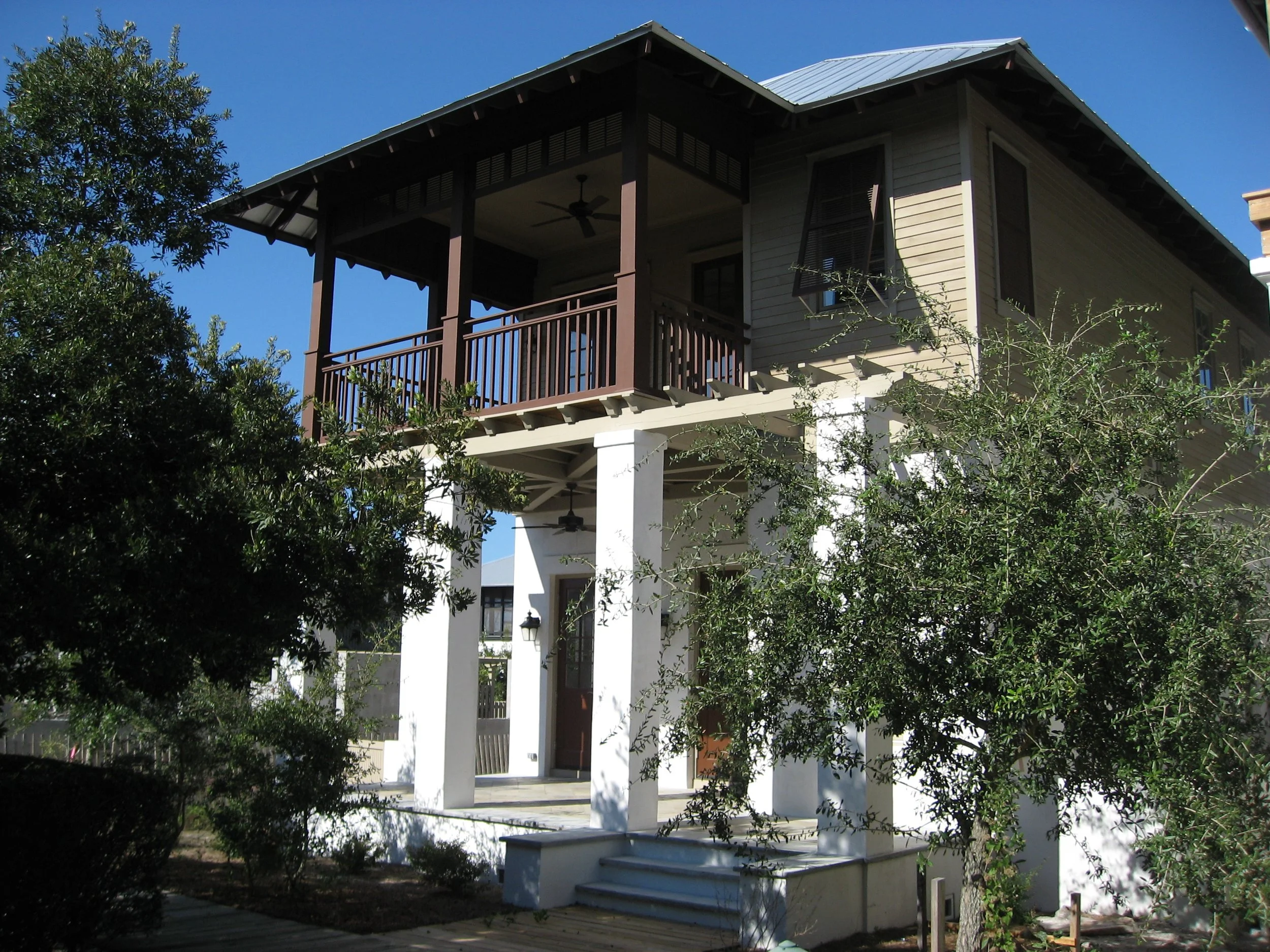 A two-story house with a balcony, white columns, and a staircase leading to the front door, surrounded by trees and greenery under a clear blue sky.