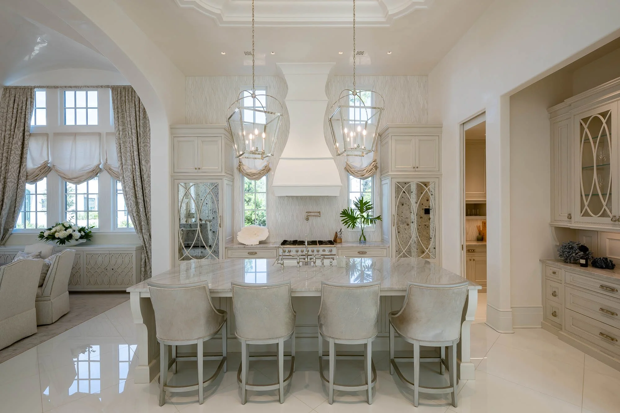 Bright, elegant kitchen with a large island, five beige upholstered chairs, white cabinetry, and two geometric pendant lights. Large windows with curtains and greenery outside.