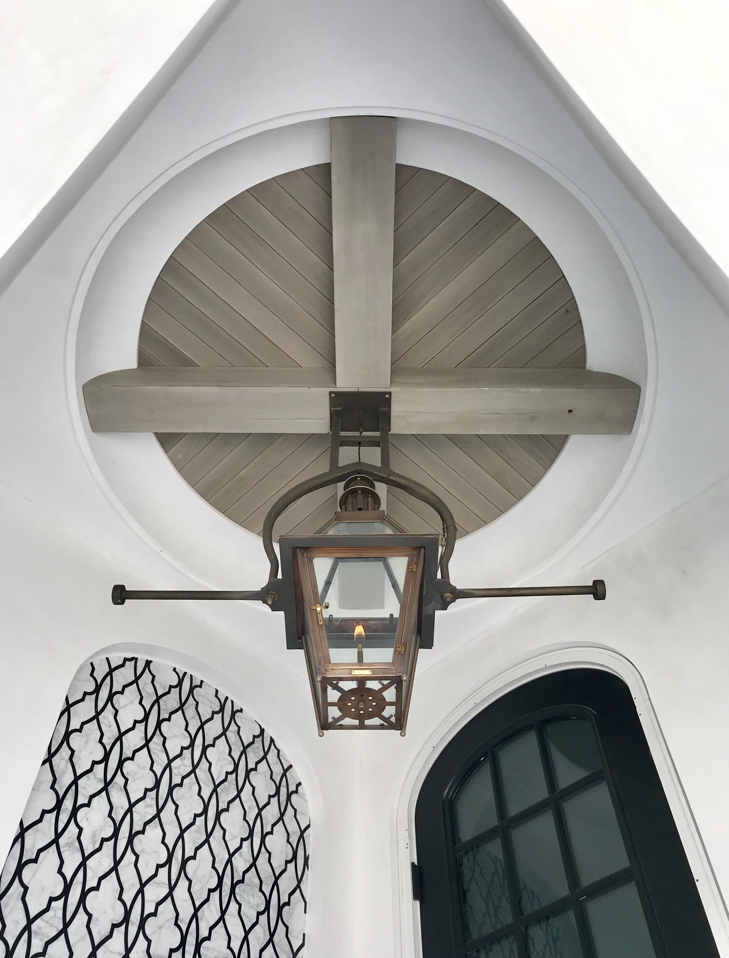 Looking up at a ceiling with a hanging lantern and a wooden round ceiling with cross beams, surrounded by white walls and decorated with black wrought iron patterned railing and a black arched window.