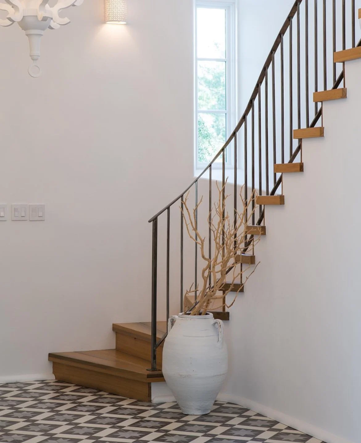 Interior of a modern house with wooden stairs, a large white vase with dried branches, patterned tile flooring, and a window allowing natural light.