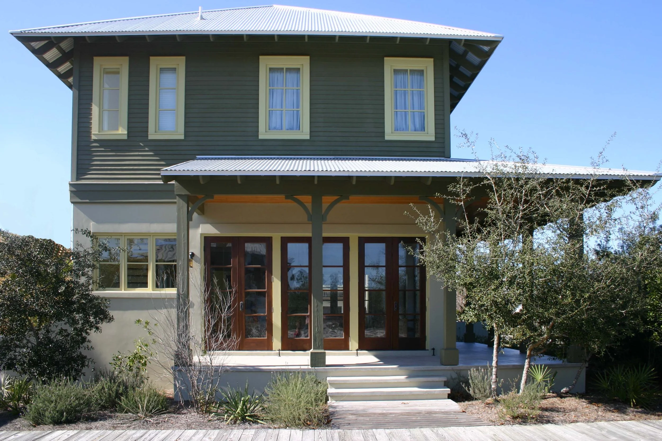 Two-story house with green siding and a white metal roof, wooden front porch with steps, and surrounding bushes and small trees.