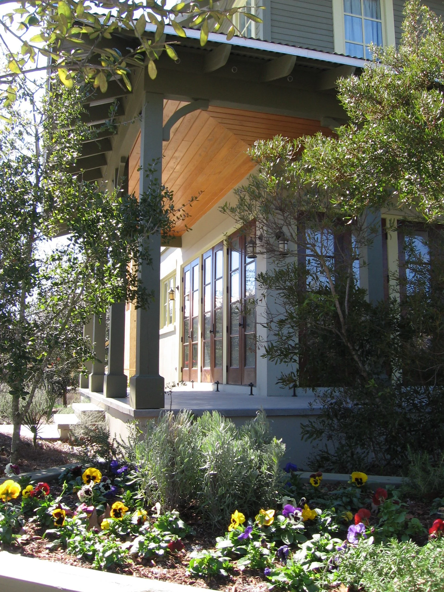 Front porch of a house with glass double doors, wooden ceiling, columns, and a garden with colorful flowers and shrubs