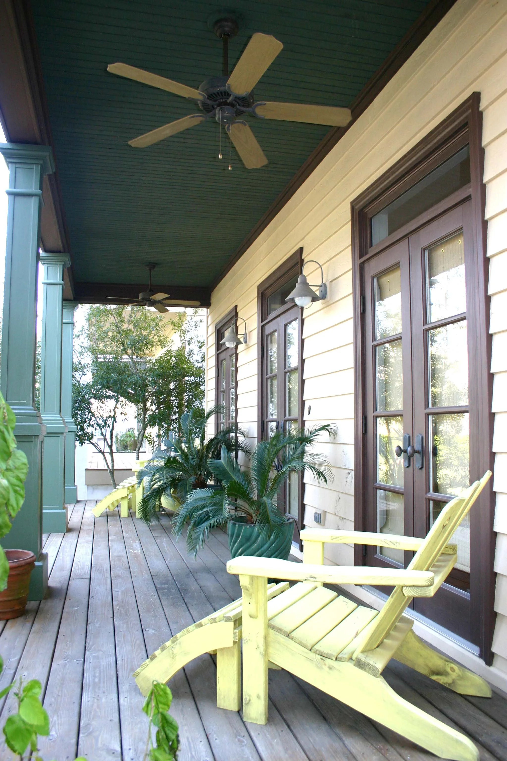 Front porch of a house with yellow siding, wooden deck, potted plants, yellow Adirondack chair, and ceiling fans with green ceiling.