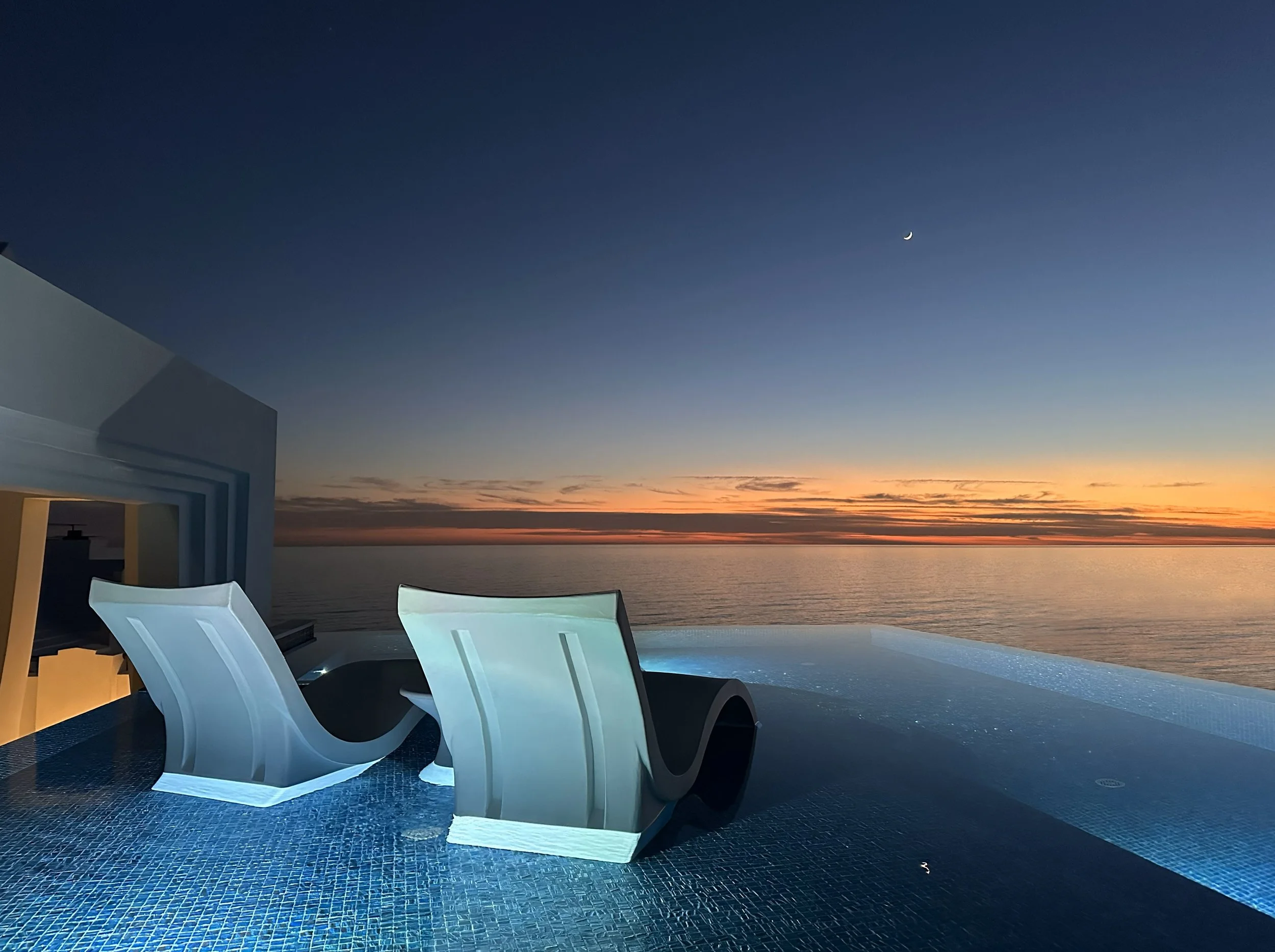 Luxury outdoor pool area with modern white lounge chairs on tiled deck, overlooking ocean during sunset with colorful sky and crescent moon.