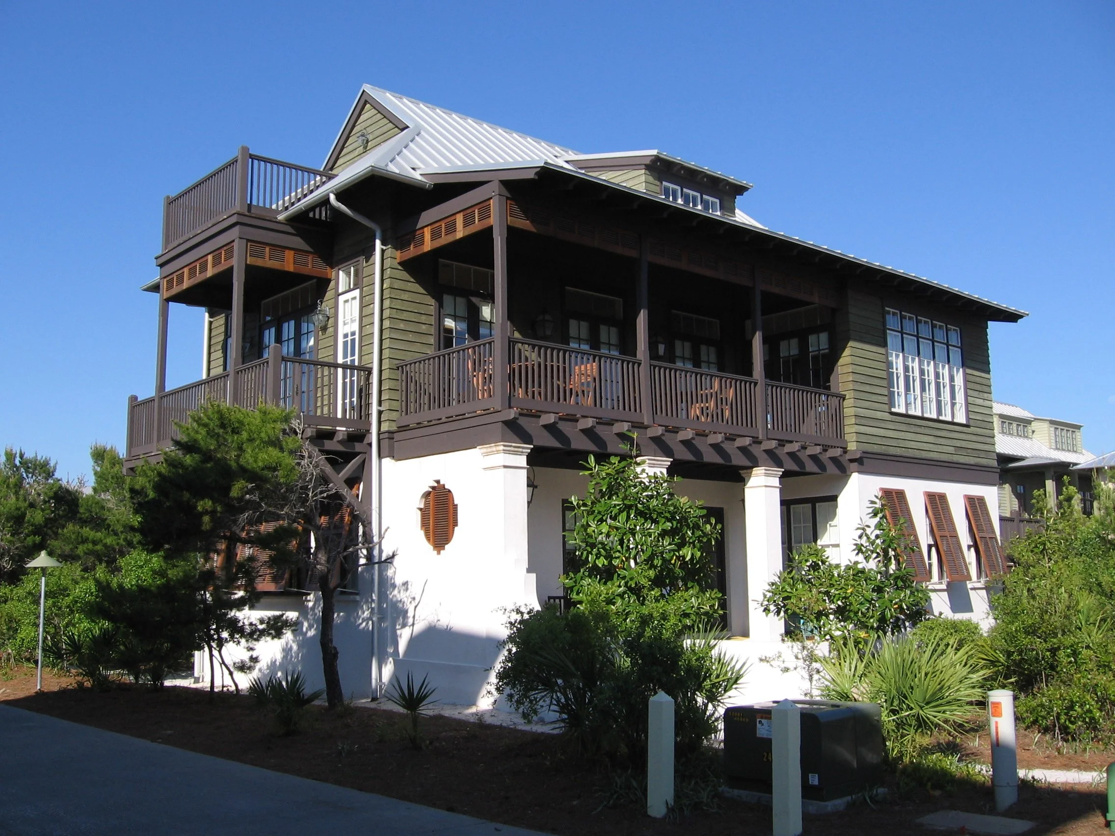 A large multi-story house with a green and white exterior, multiple decks with wooden railings, large windows, and a sloped metal roof, surrounded by bushes and trees.