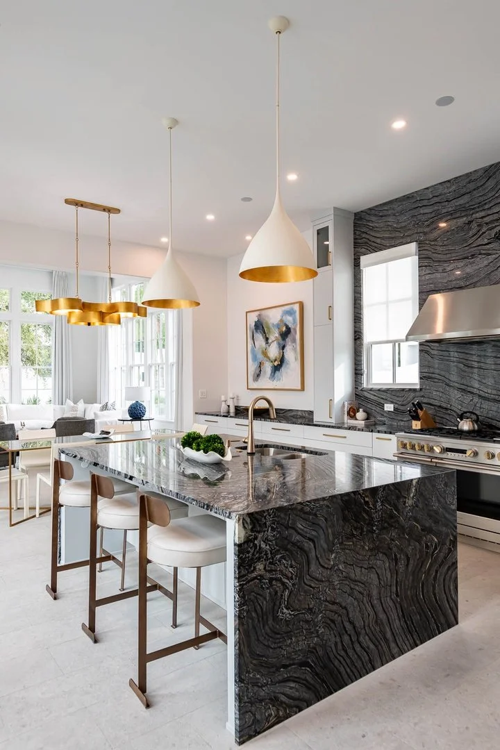 Modern kitchen with a dark patterned waterfall island, white cabinetry, and gold pendant lights. There are windows and artwork in the background.
