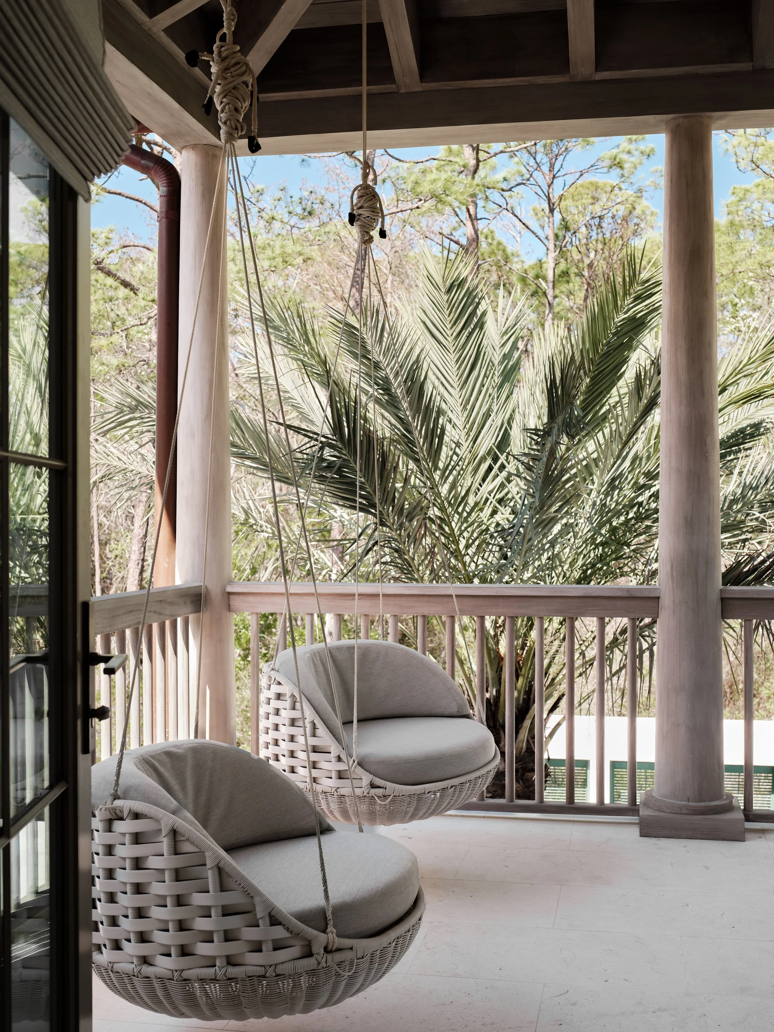 Balcony with two hanging rattan swing chairs with cushions, overlooking lush green trees and palm plants.
