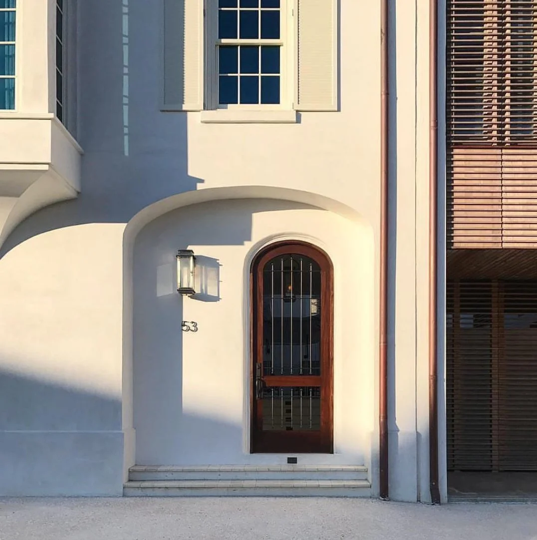 Front door of a modern house with white walls, wooden door with metal bars, steps, window shutters, and a wall-mounted light fixture.