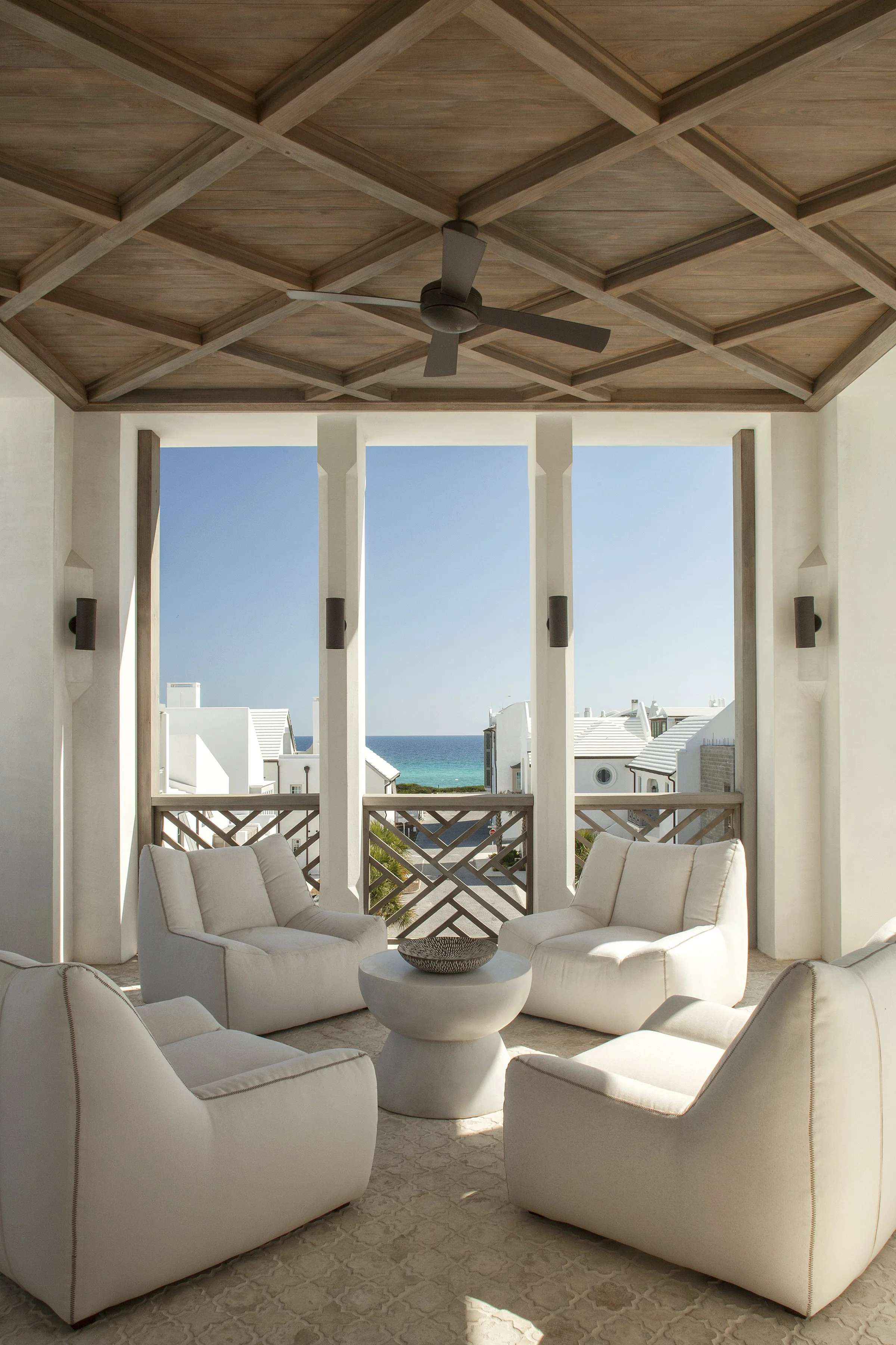 A covered balcony with four white armchairs arranged around a small white round table, overlooking a seaside neighborhood with white houses and a view of the ocean and clear blue sky.