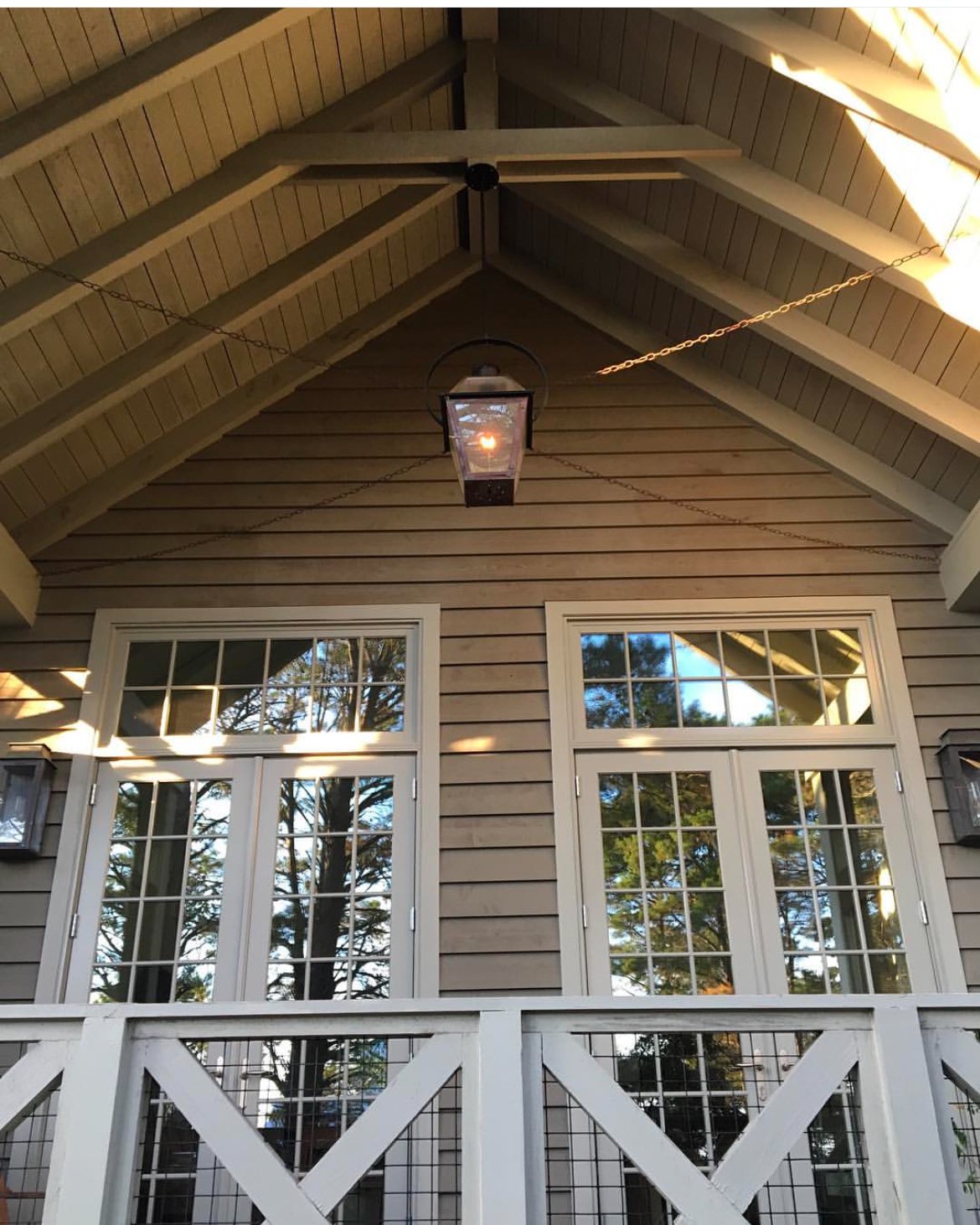 View of a house porch ceiling with a hanging lantern light, two sets of white framed windows, a white porch railing, and reflections of trees in the windows.