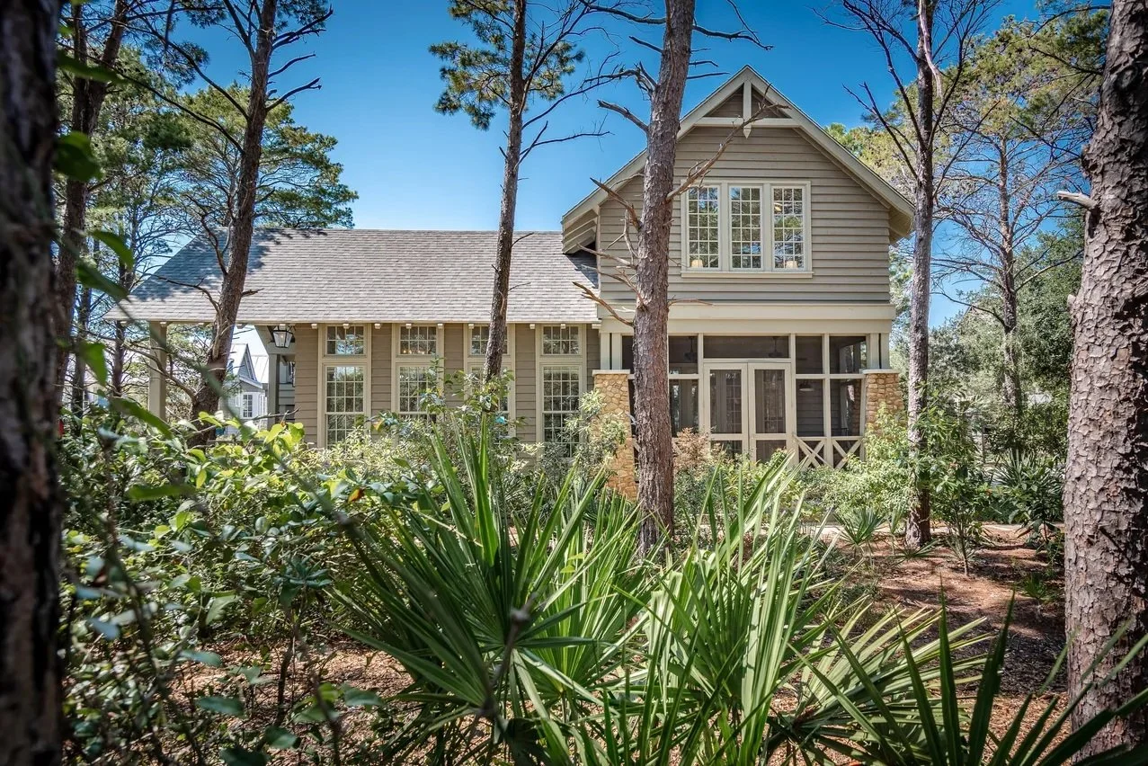 A two-story house with a beige exterior, large windows, and a screened porch surrounded by tall trees and greenery on a sunny day.