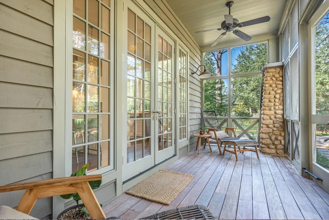 Sunlit screened porch with wooden deck, glass double doors, ceiling fan, stone pillar, outdoor chairs, and trees visible outside.