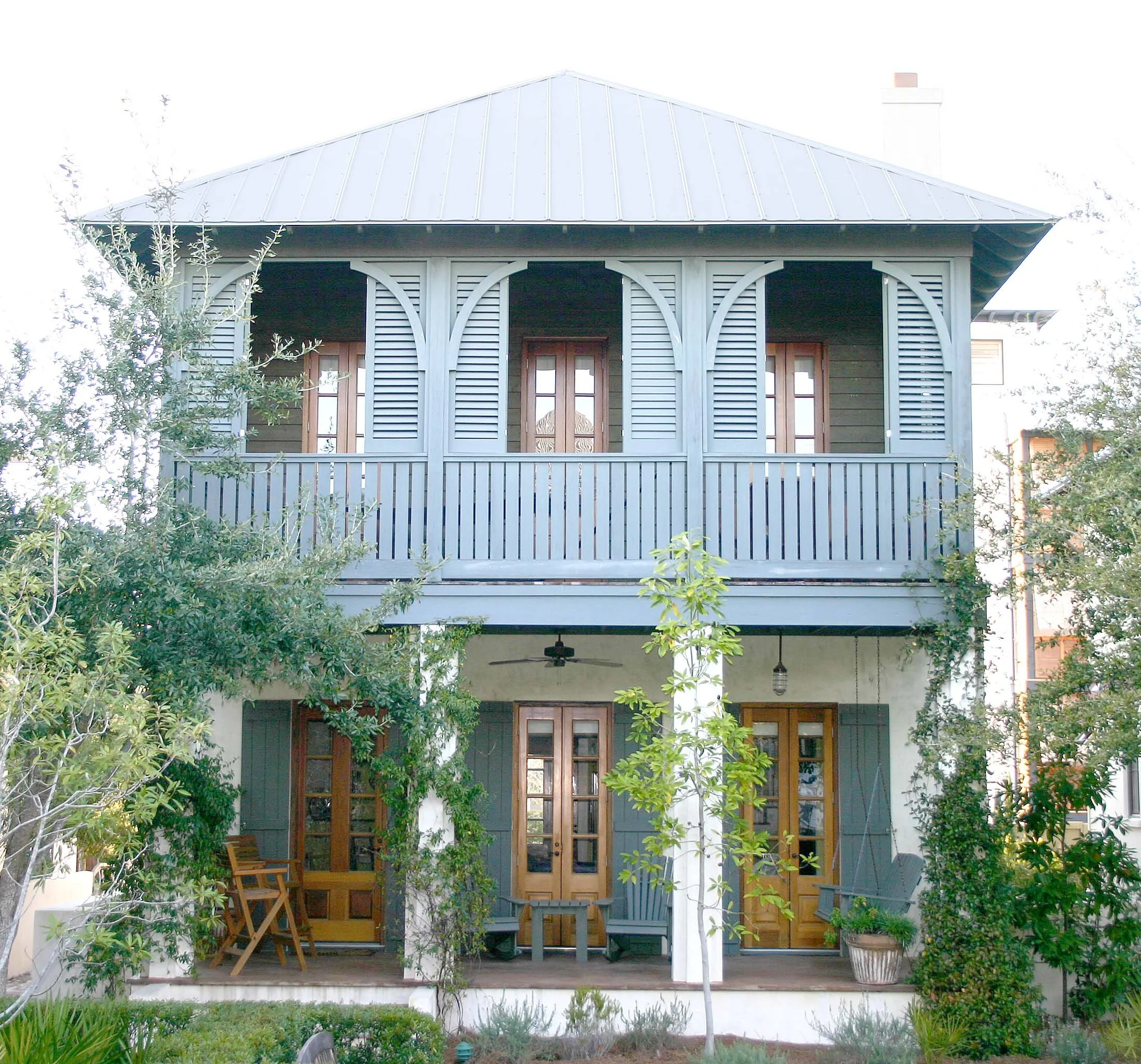 Two-story house with a gray exterior, wooden doors and window shutters, a balcony on the second floor with blue railings, surrounded by greenery and trees.