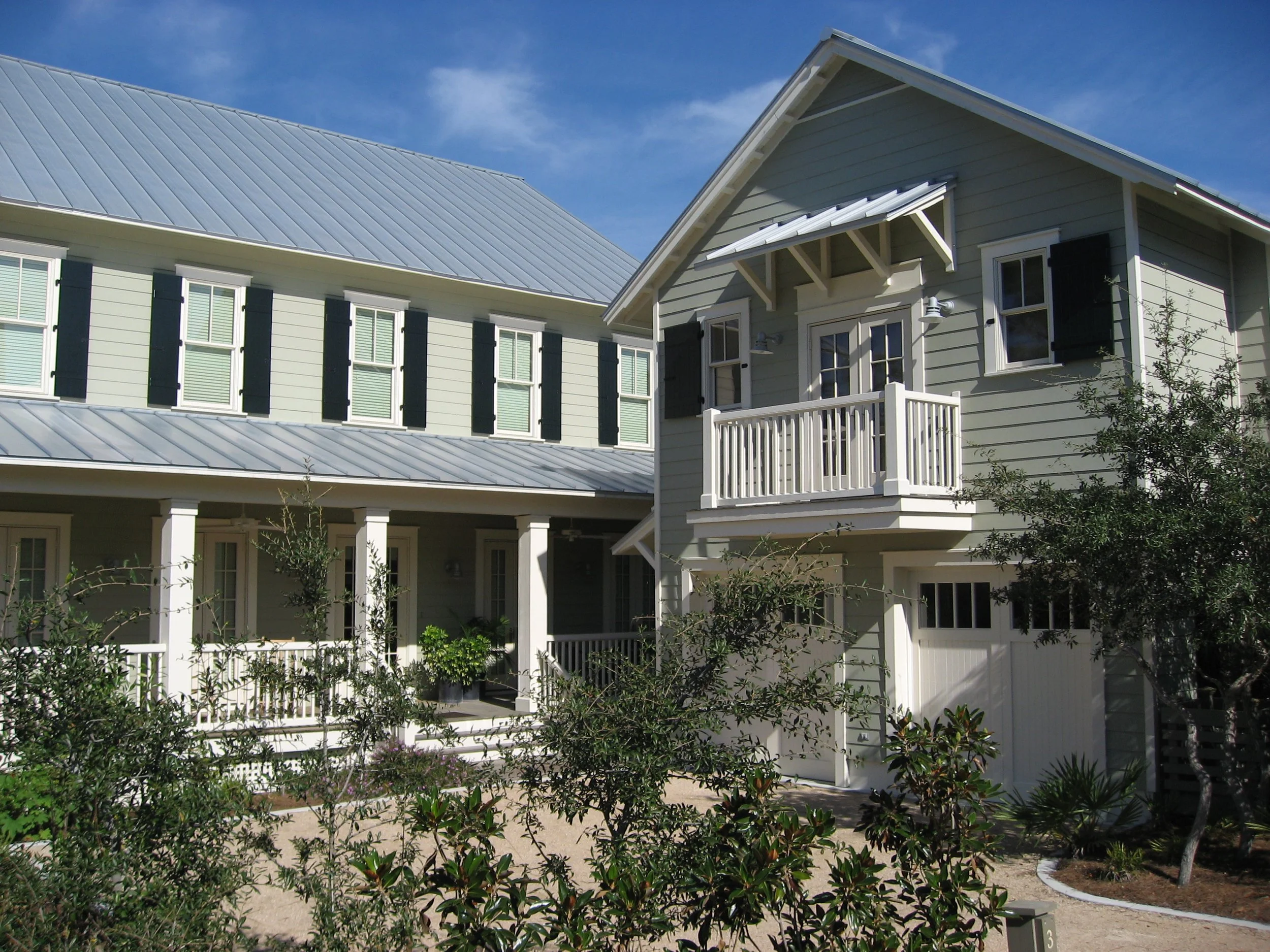 A two-story house with a light grey exterior and white trim, featuring a small balcony, green shutters, and a metal roof, surrounded by shrubs and plants under a blue sky.