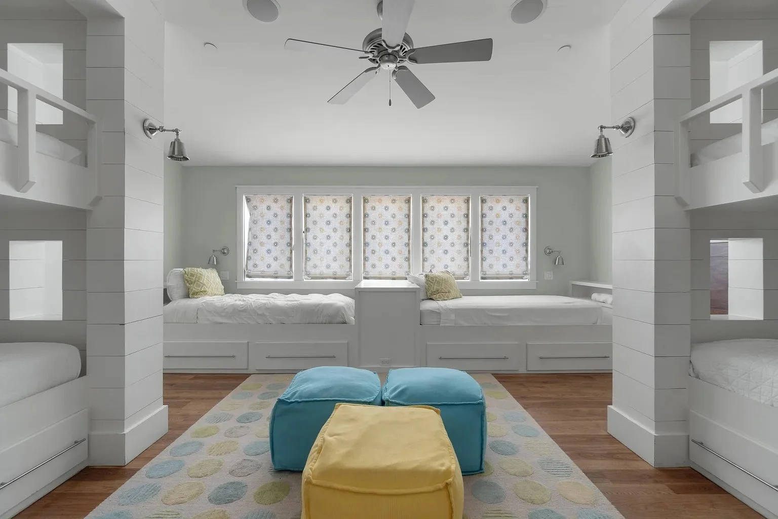 Upside-down bedroom with four beds beneath a row of five windows with patterned curtains, ceiling fan, hardwood flooring, and colorful poufs and rug.