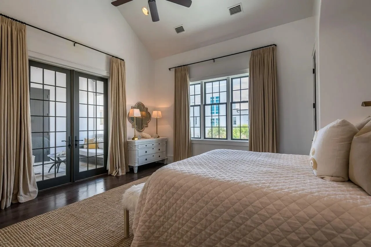 A bedroom with a large bed, beige quilt, and pillows, a white dresser with lamps, large windows with beige curtains, wooden floor, and a ceiling fan.