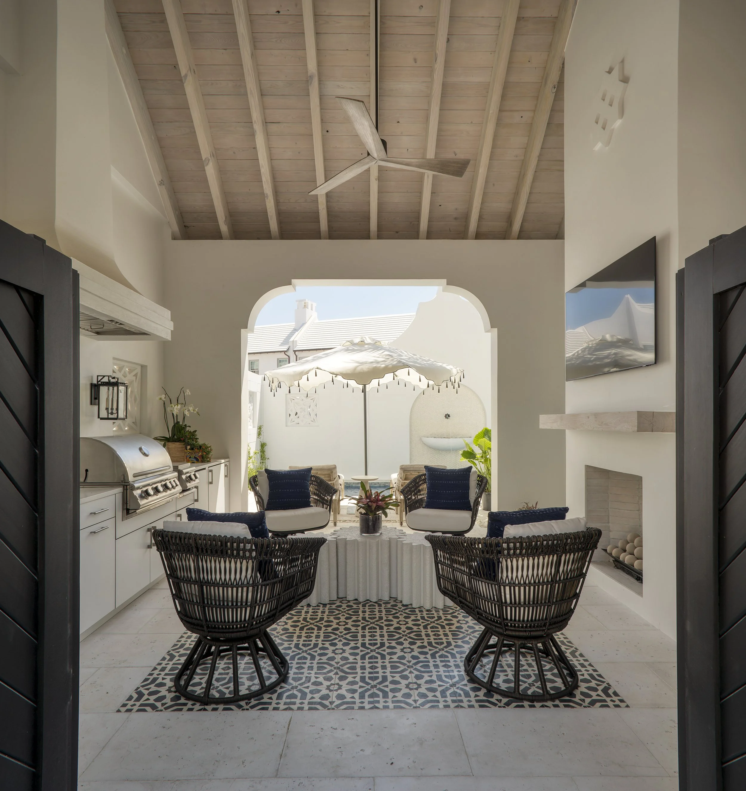 Outdoor patio with four black woven chairs around a white ruffled table, shaded by a large white umbrella, with a built-in grill, plants, and a water feature in the background.