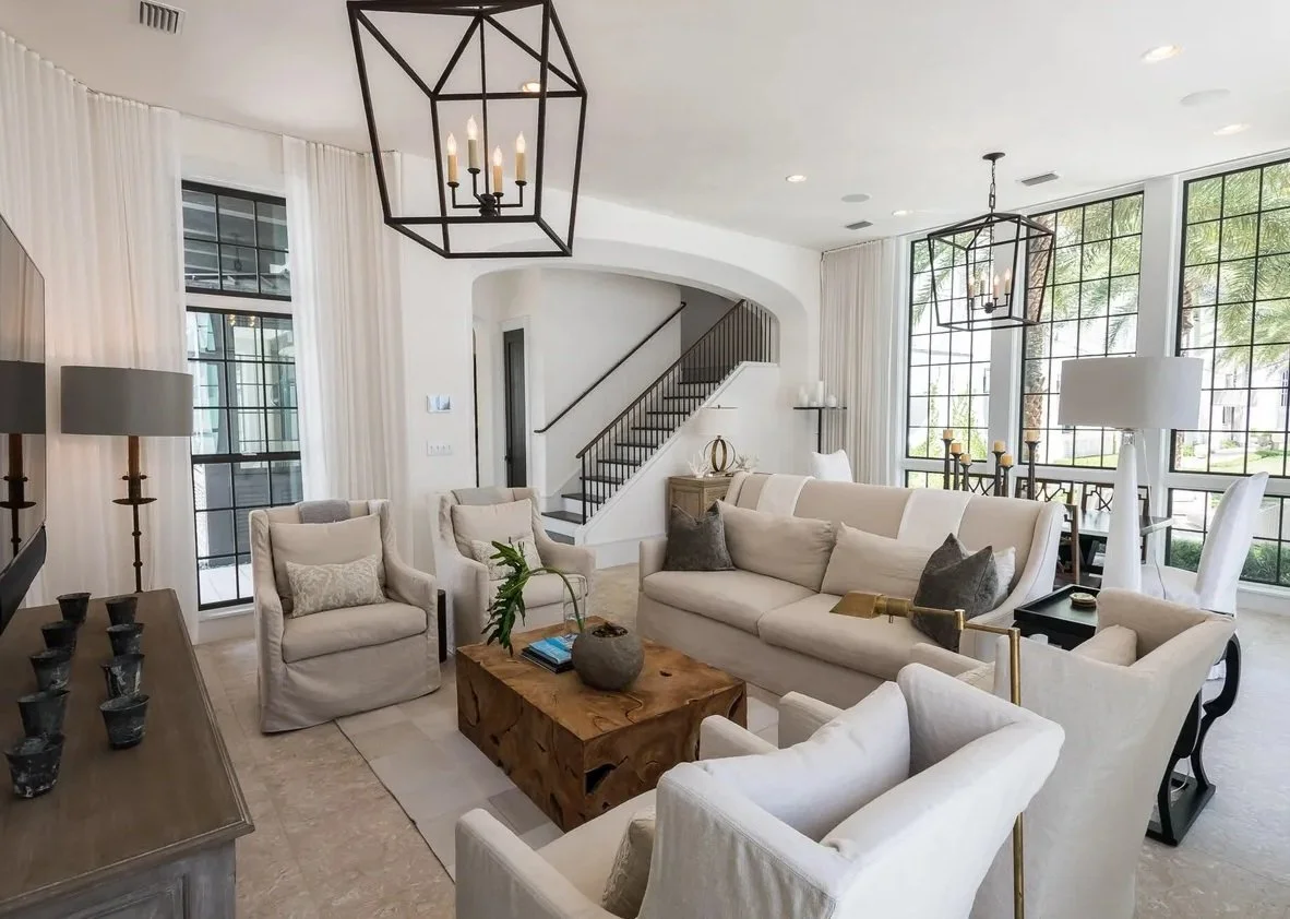 Bright modern living room with white furniture, large window, black metal light fixtures, and a wooden coffee table.