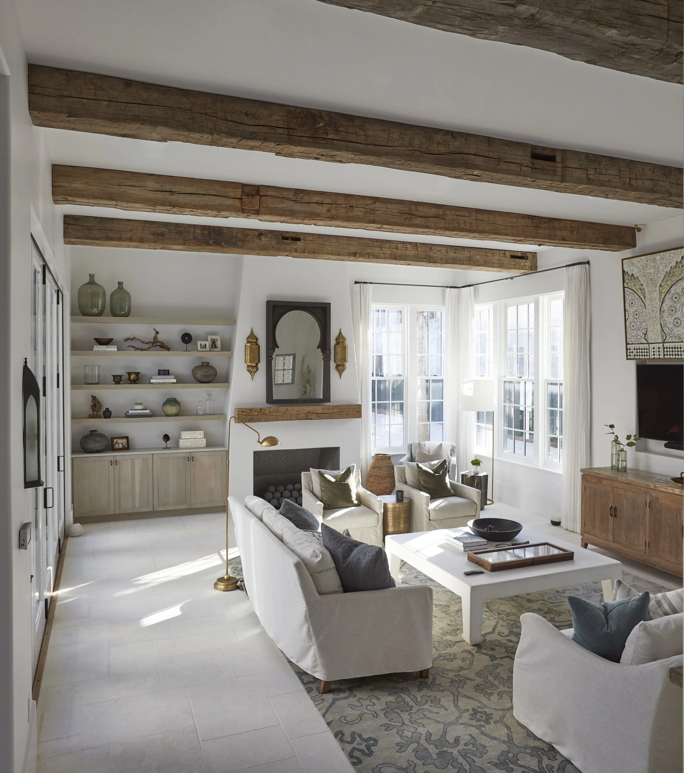 Living room with white sofas, armchairs, wooden beams on ceiling, large windows with white curtains, and a fireplace