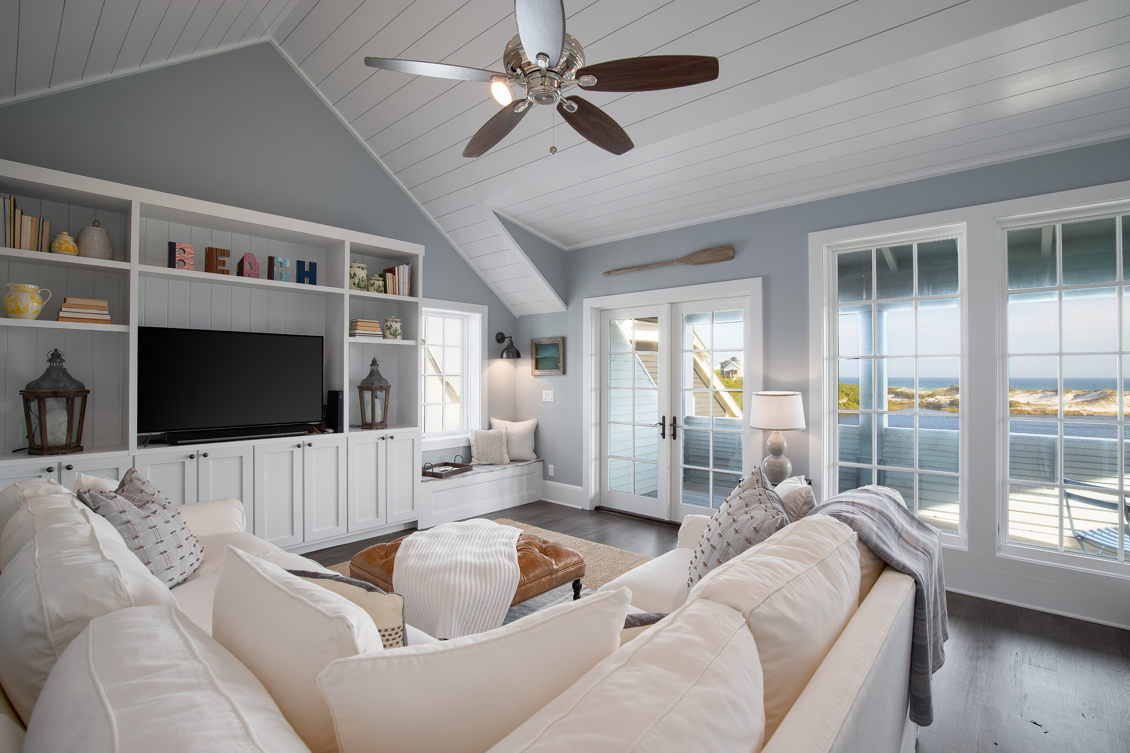 Bright coastal living room with white sectional sofa, dark hardwood floors, large windows and glass doors showing ocean views, built-in white shelving unit with TV, decorative items, and books, ceiling fan with wooden blades, and a night bench under 