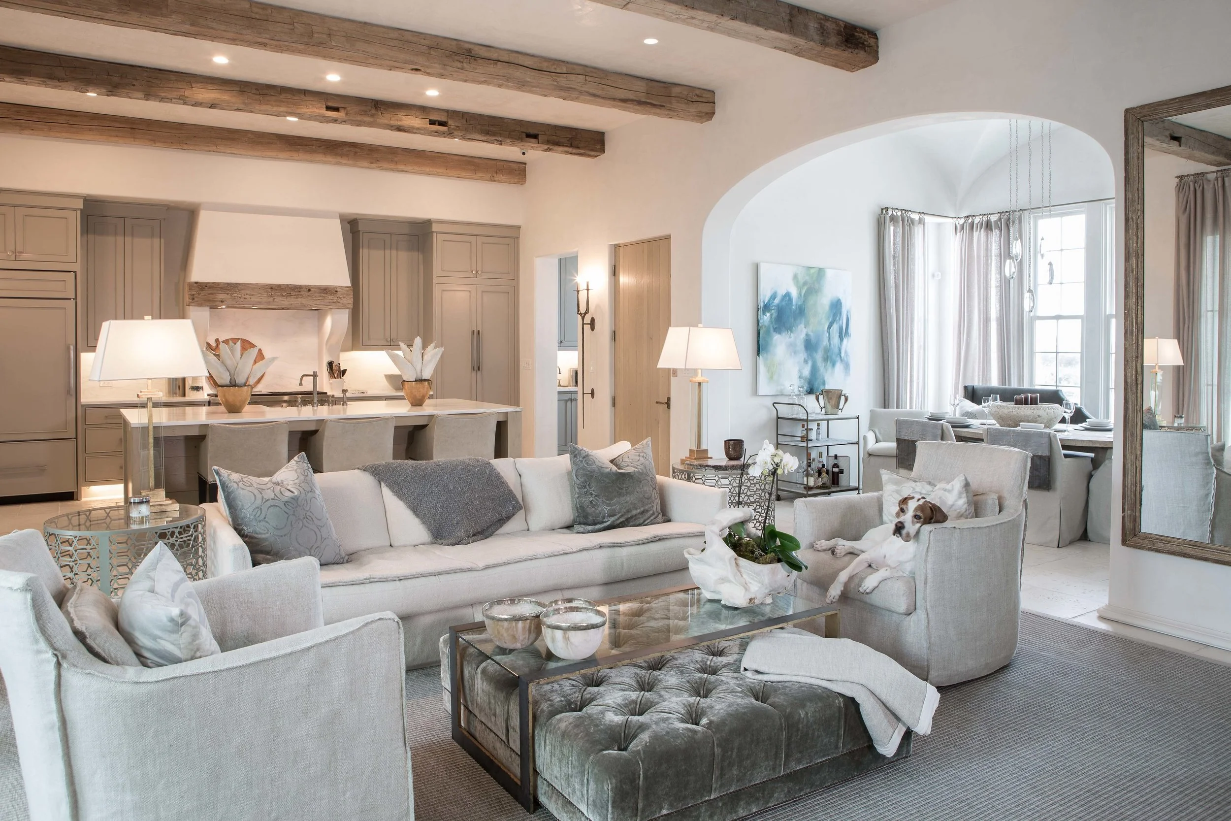 Bright and spacious living room with neutral tones, velvet and fabric sofas, a tufted ottoman, artwork, and a dog relaxing on one of the chairs. Open kitchen with an island and modern cabinetry in the background.