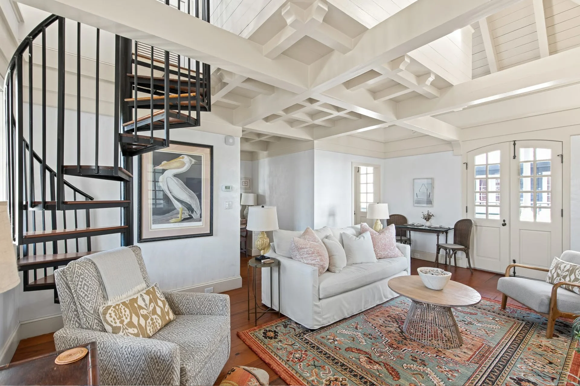 Living room with white walls, a white ceiling with exposed beams, a spiral staircase with black railing, a white sofa with pink and beige pillows, a patterned armchair, a wooden coffee table, a colorful area rug, and large windows with white trim.