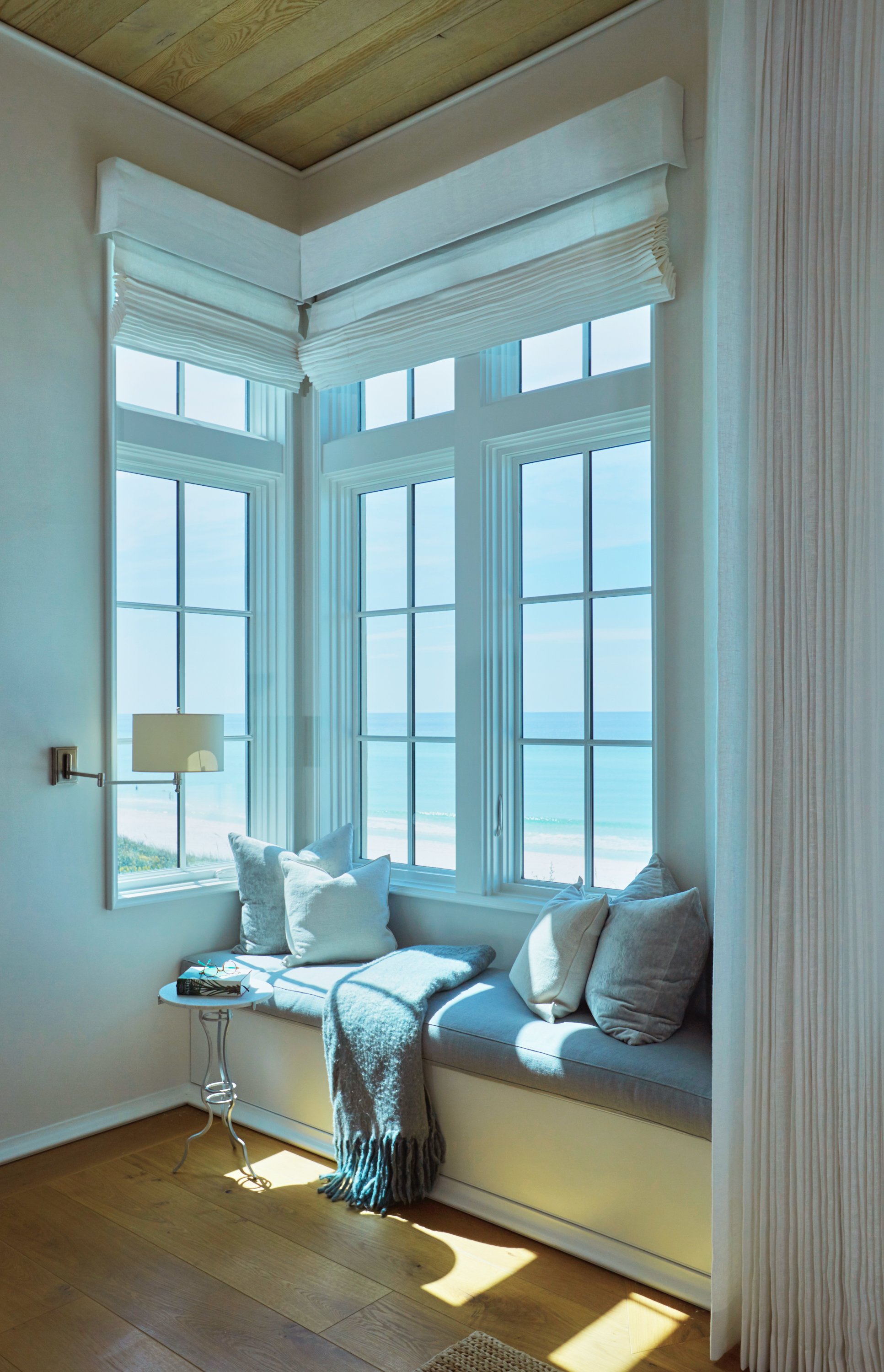 Bright beachside room with large windows showing ocean view, window seat with cushions, throw blanket, and small side table with books and glasses.