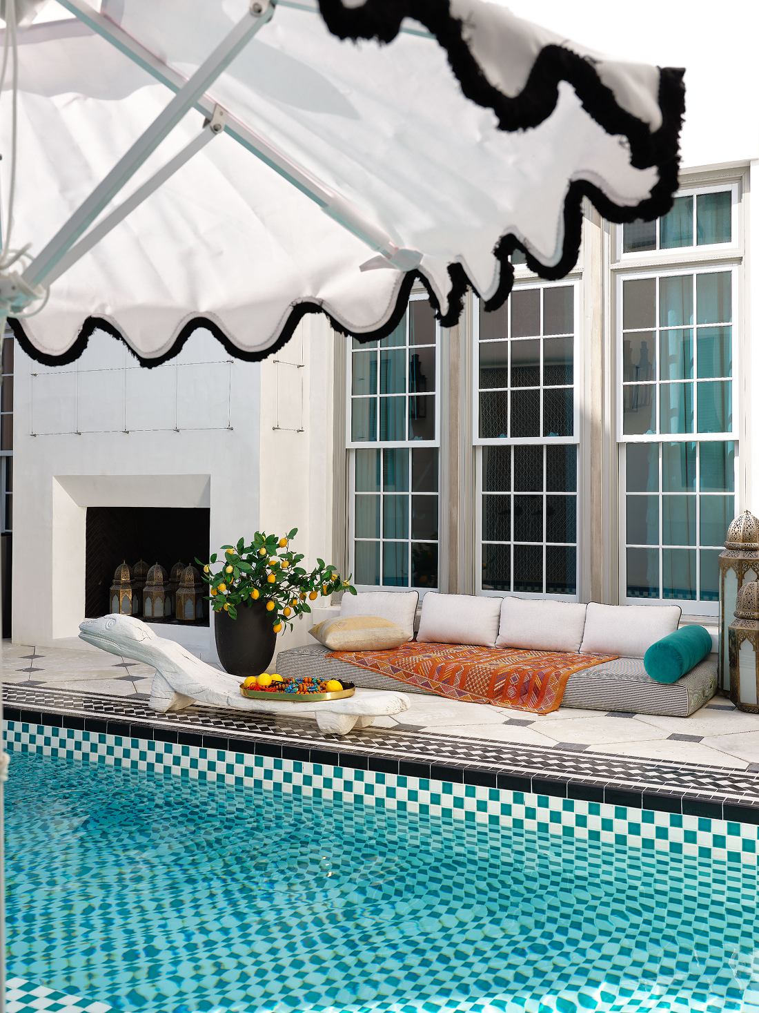 Poolside patio with a white lounge chair, a potted lemon tree, cushions on a built-in bench, amber and teal pillows, a patterned blanket, lanterns, and an umbrella overhead.