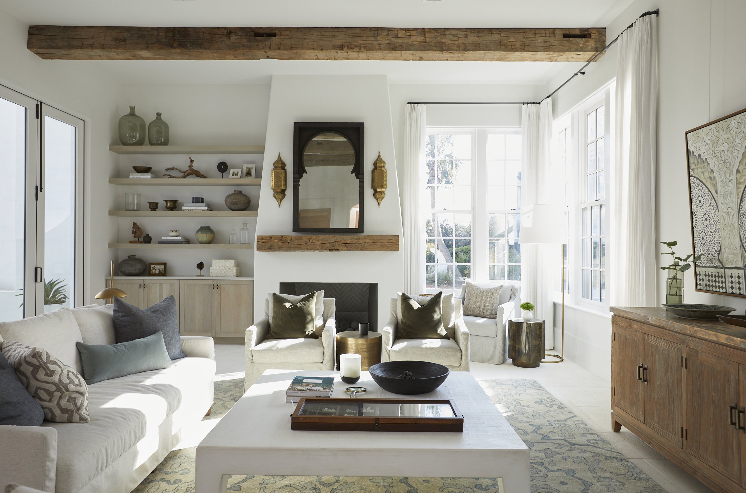 Bright living room with white sofas, armchairs, and a wooden sideboard, featuring decorative pillows, vases, and artwork, with large windows and built-in shelves.