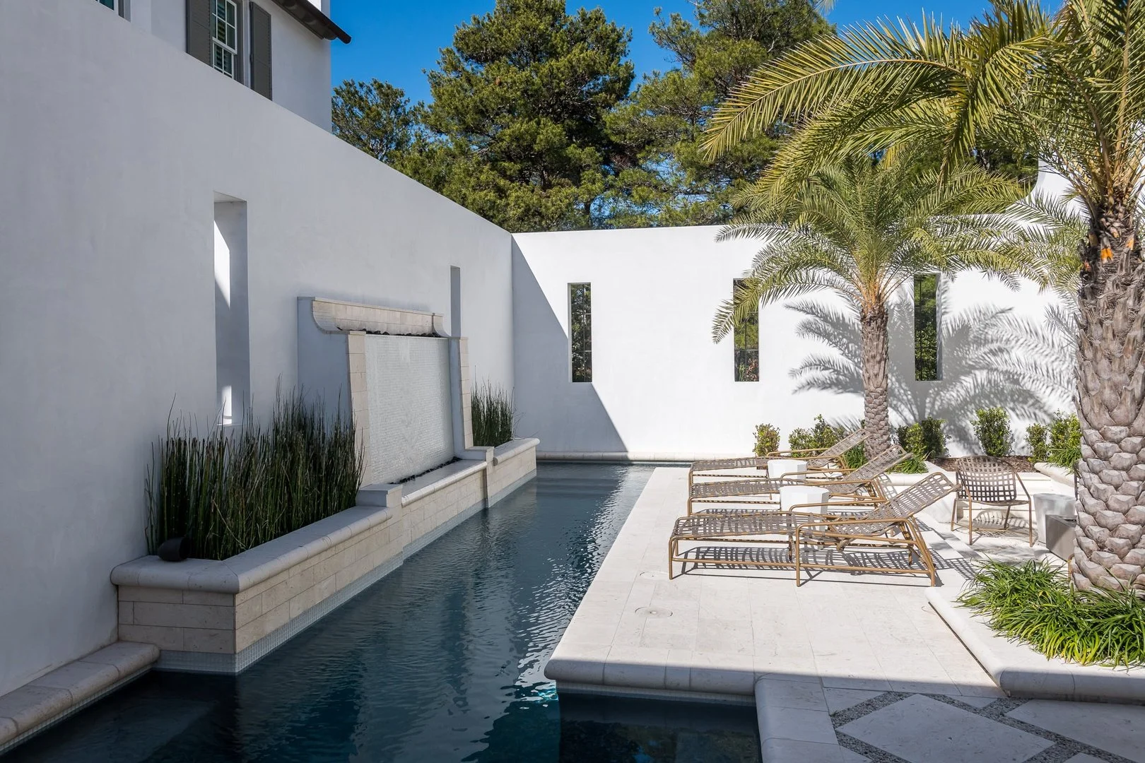 Luxury outdoor pool area with white walls, palm trees, and lounge chairs under blue sky.